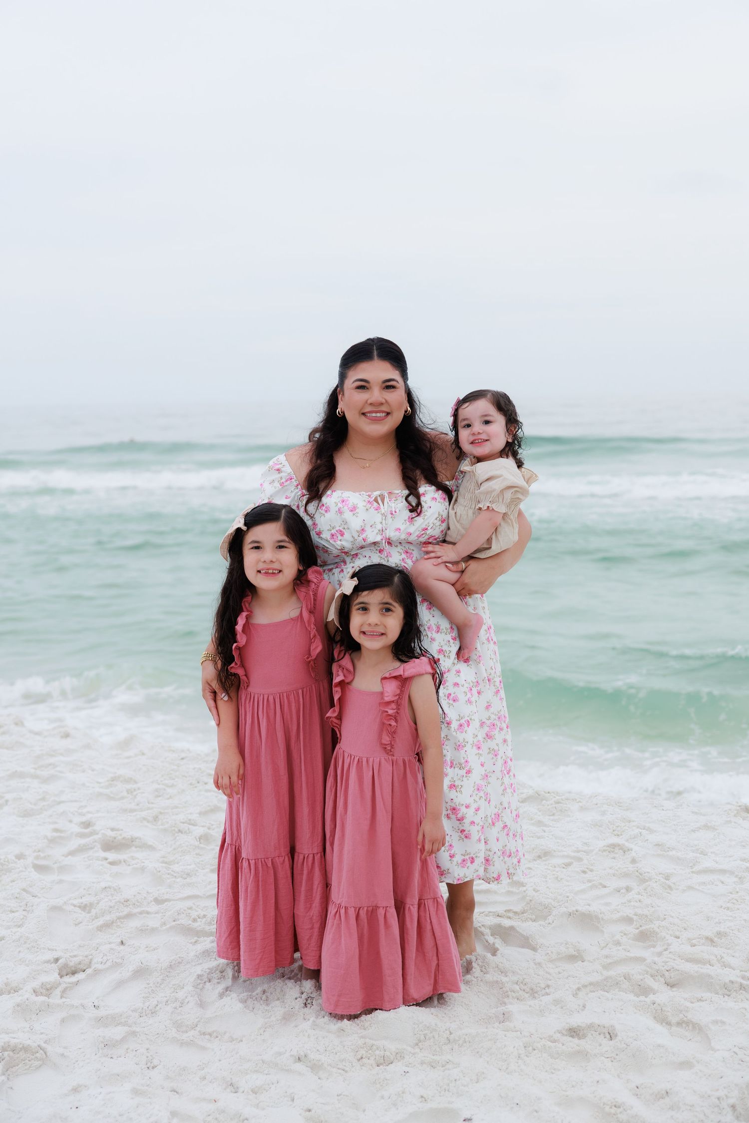 A mother and three young daughters pose together on a beach, wearing pink dresses, with turquoise ocean waves behind them.