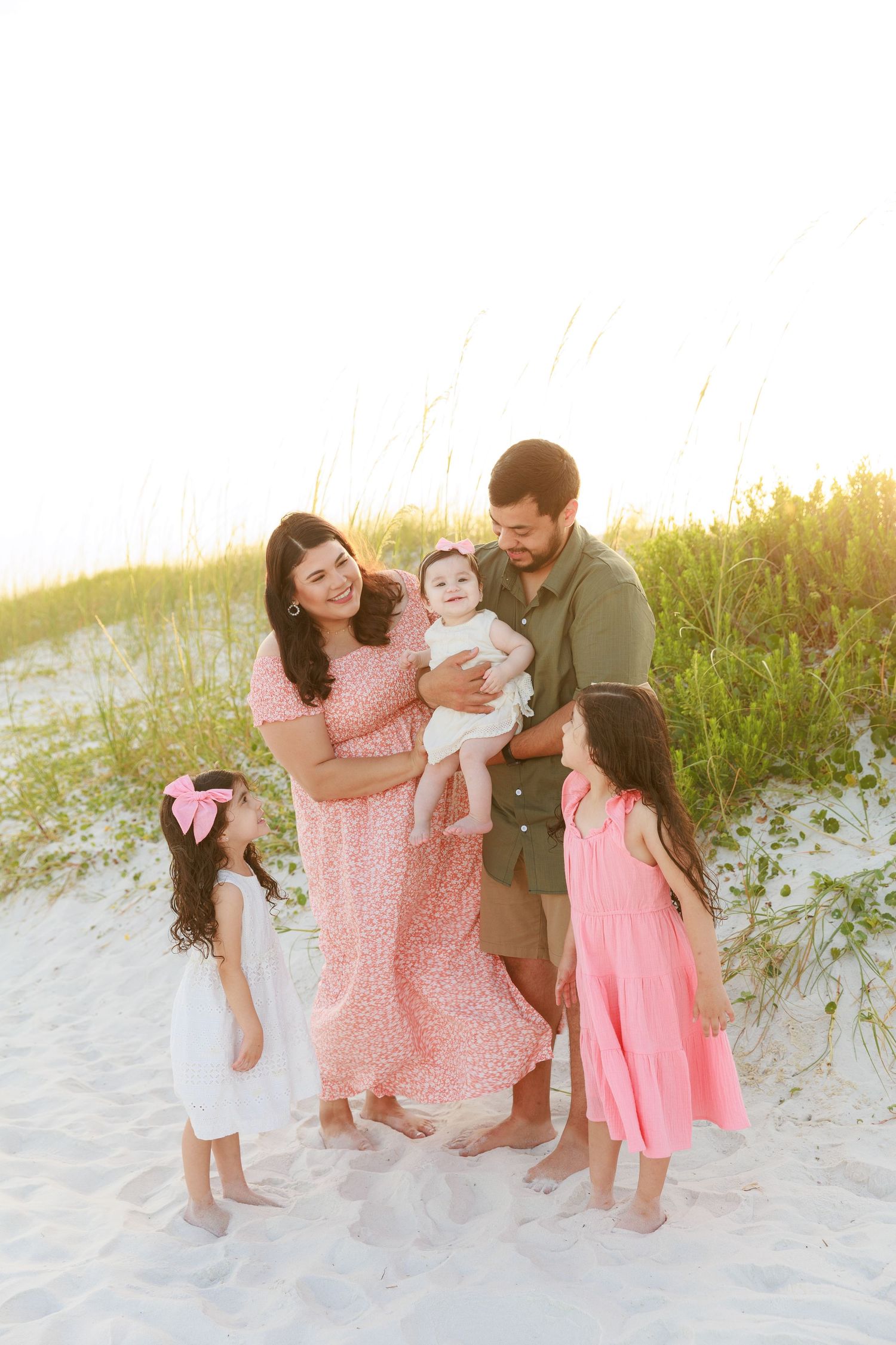 Family of five poses on sandy beach dunes at sunset, parents holding baby while two young girls wear pink and white dresses.