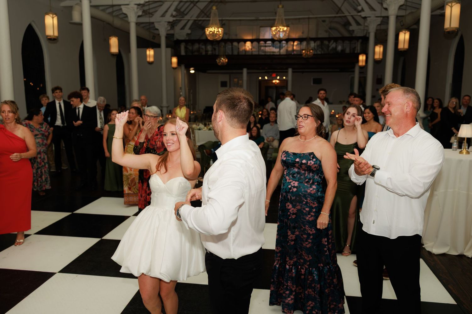 Guests dancing joyfully at an elegant wedding reception on a black and white checkered dance floor in a grand venue.