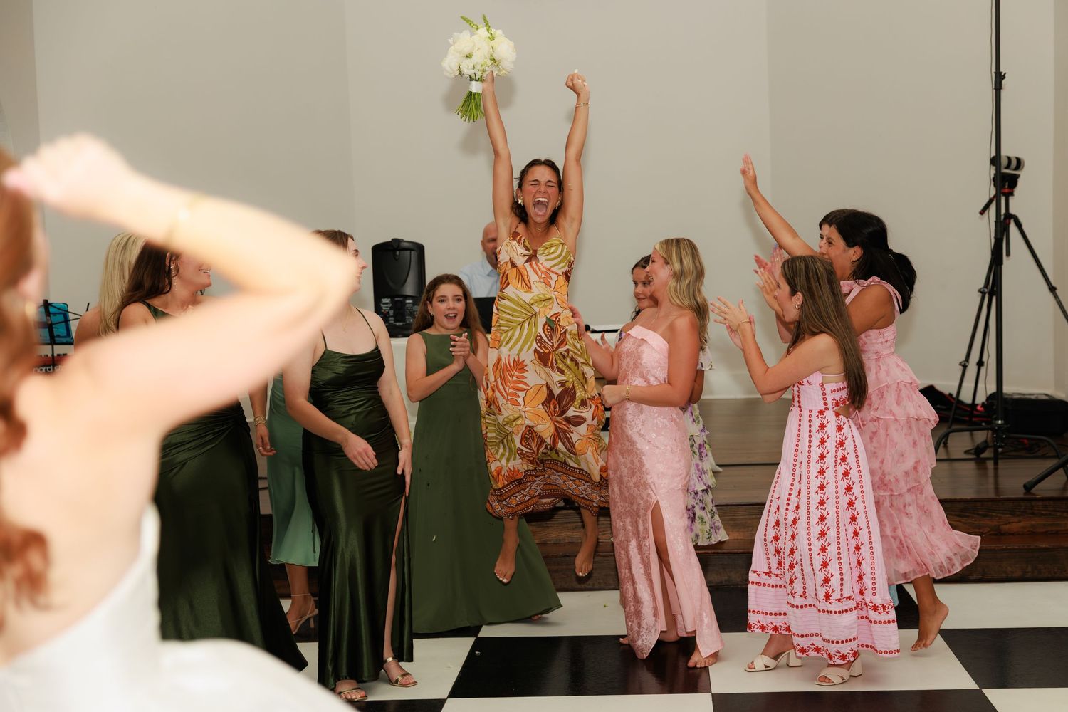 Excited woman jumps while catching bridal bouquet surrounded by cheering guests on black and white dance floor.