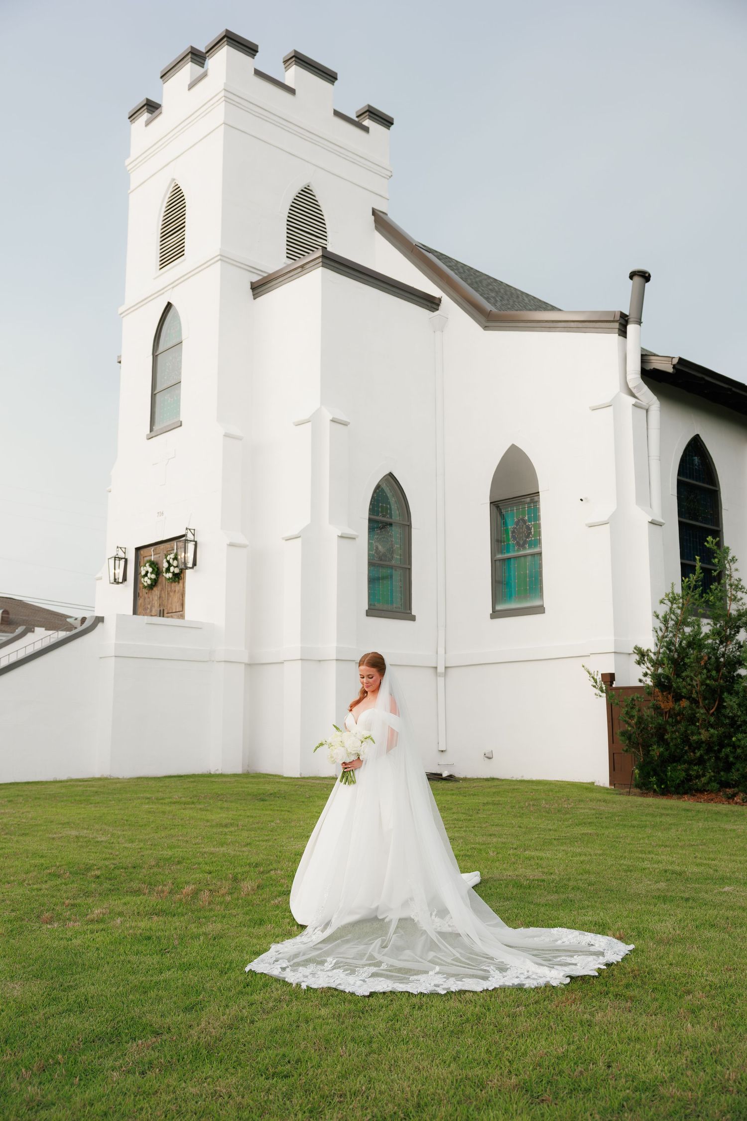 Bride in elegant white gown with long train poses in front of white Gothic-style church on green lawn.