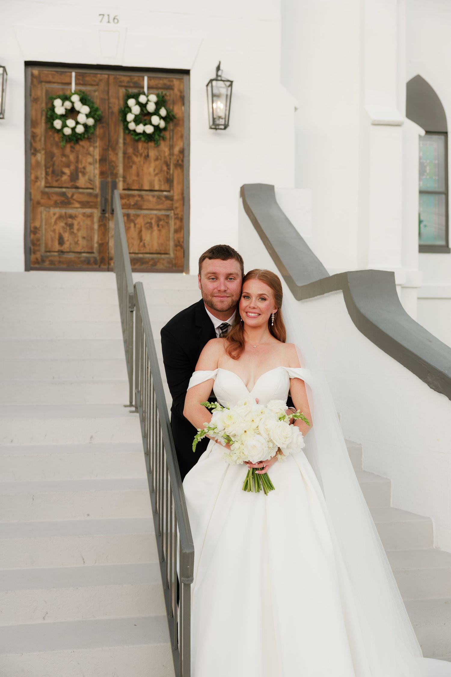 Bride and groom pose together on white church steps, bride holding white bouquet in elegant off-shoulder gown.