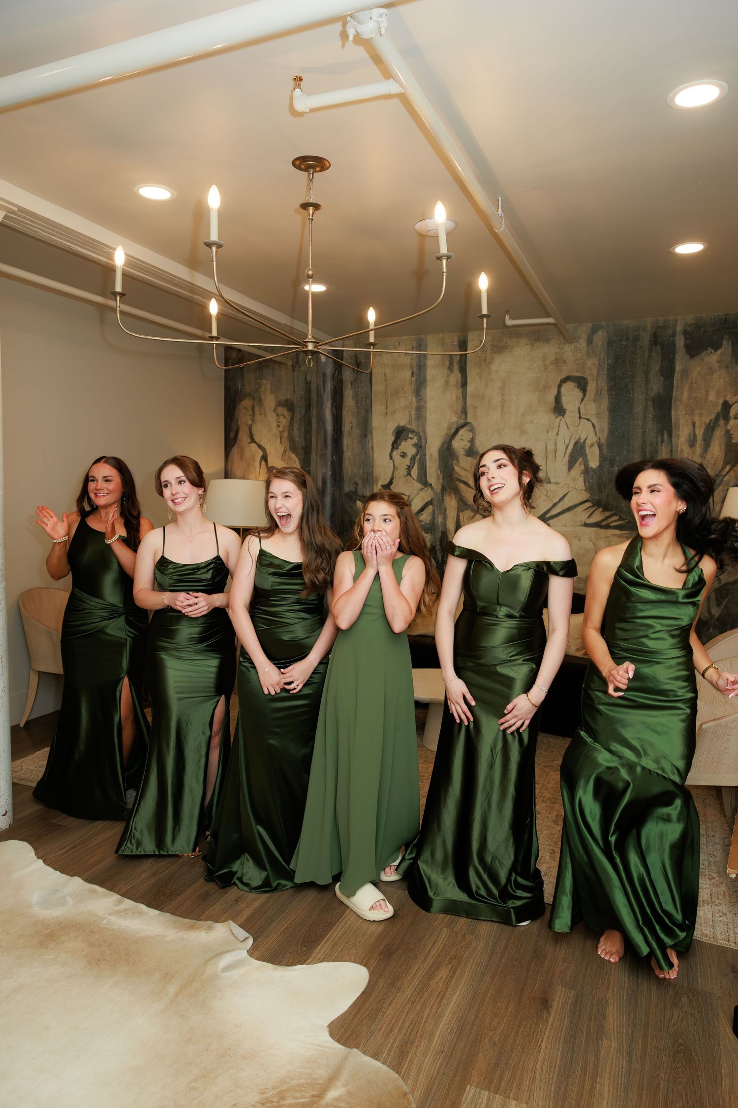 Six women in emerald green satin bridesmaid dresses react with joy and surprise in an elegant room with chandelier.