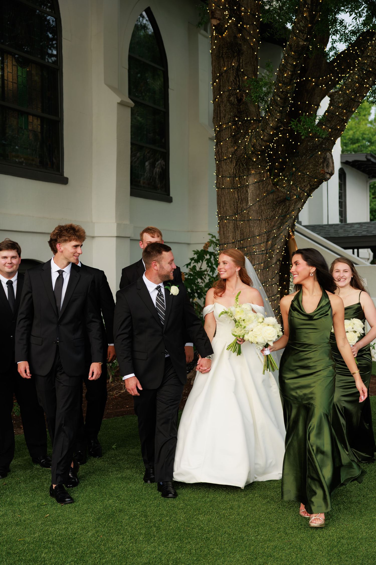 Bride and groom walk hand-in-hand with wedding party in black suits and green gowns outside a white venue.