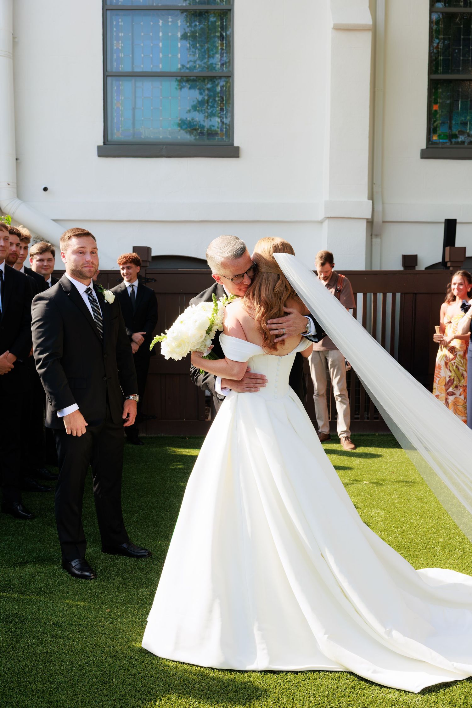 Bride in white gown embraces guest outdoors at wedding ceremony while groom in black suit watches nearby.