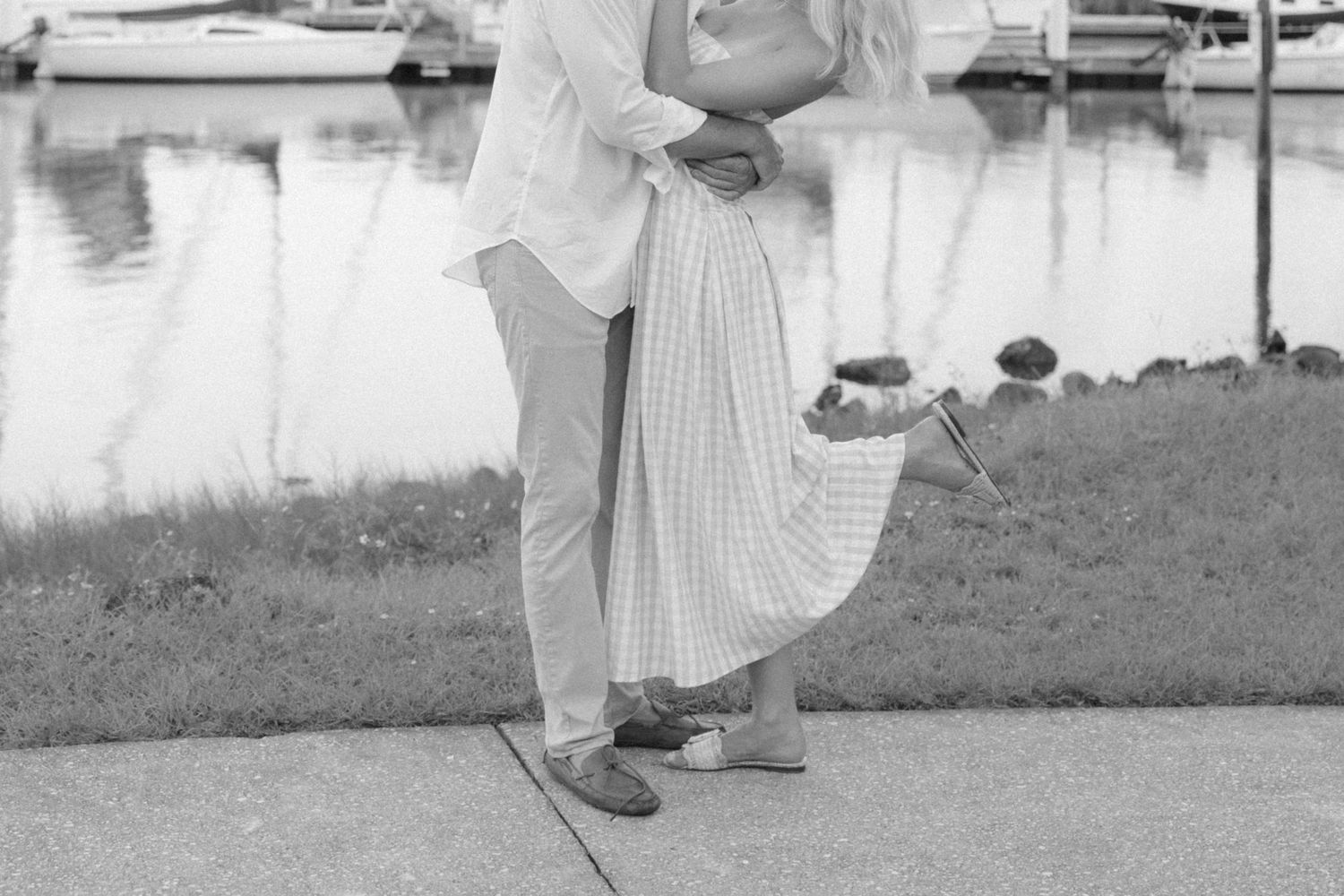 Black and white photo of a couple embracing near a marina waterfront, woman in pleated skirt and man in blazer.
