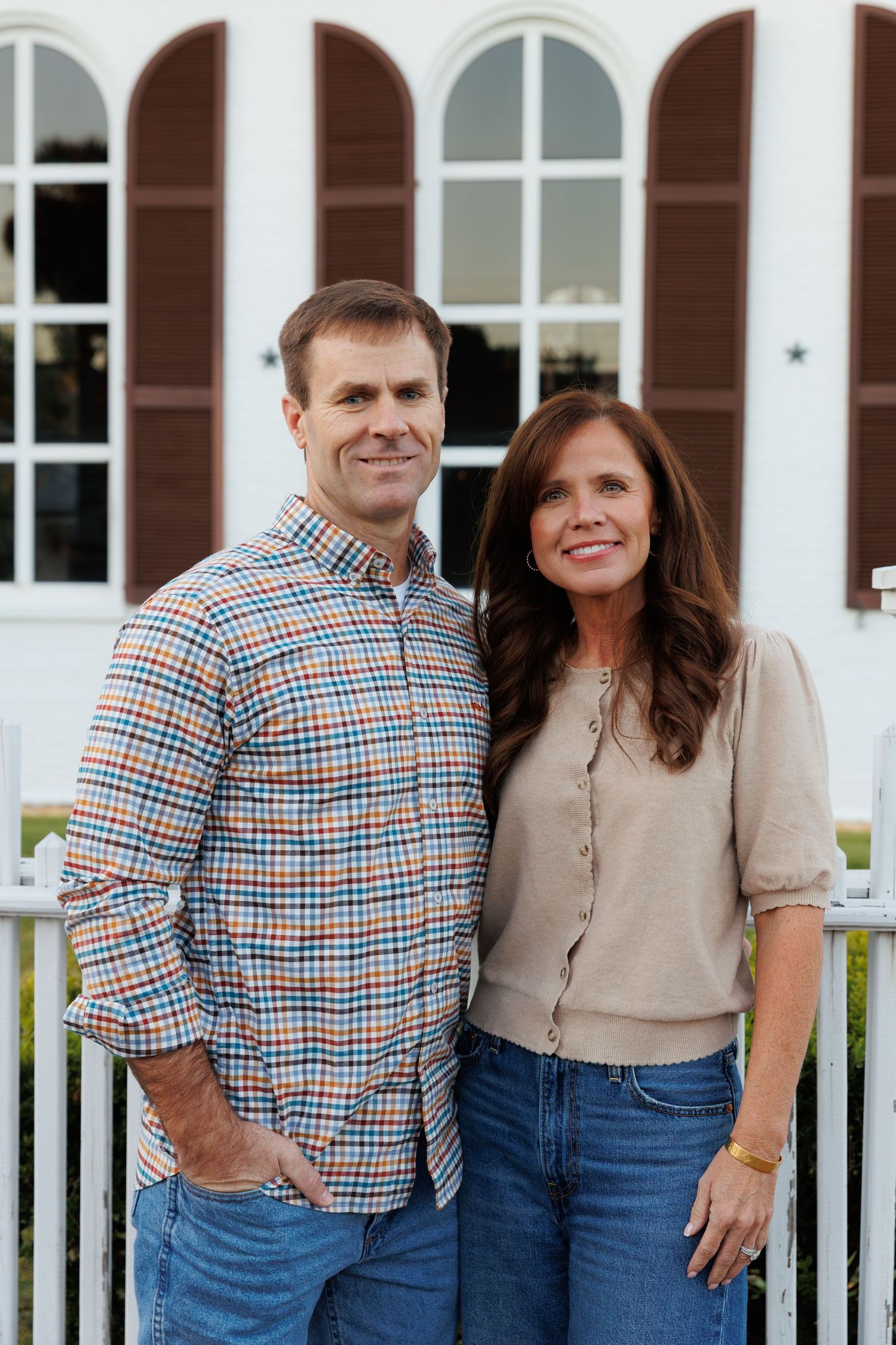 Smiling couple poses together in front of a white building with brown shutters and a white picket fence.