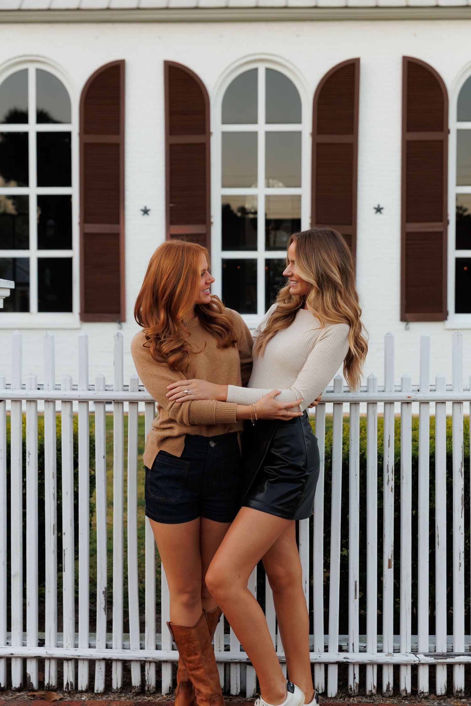 Two women in casual fall outfits embrace and smile at each other by a white picket fence.