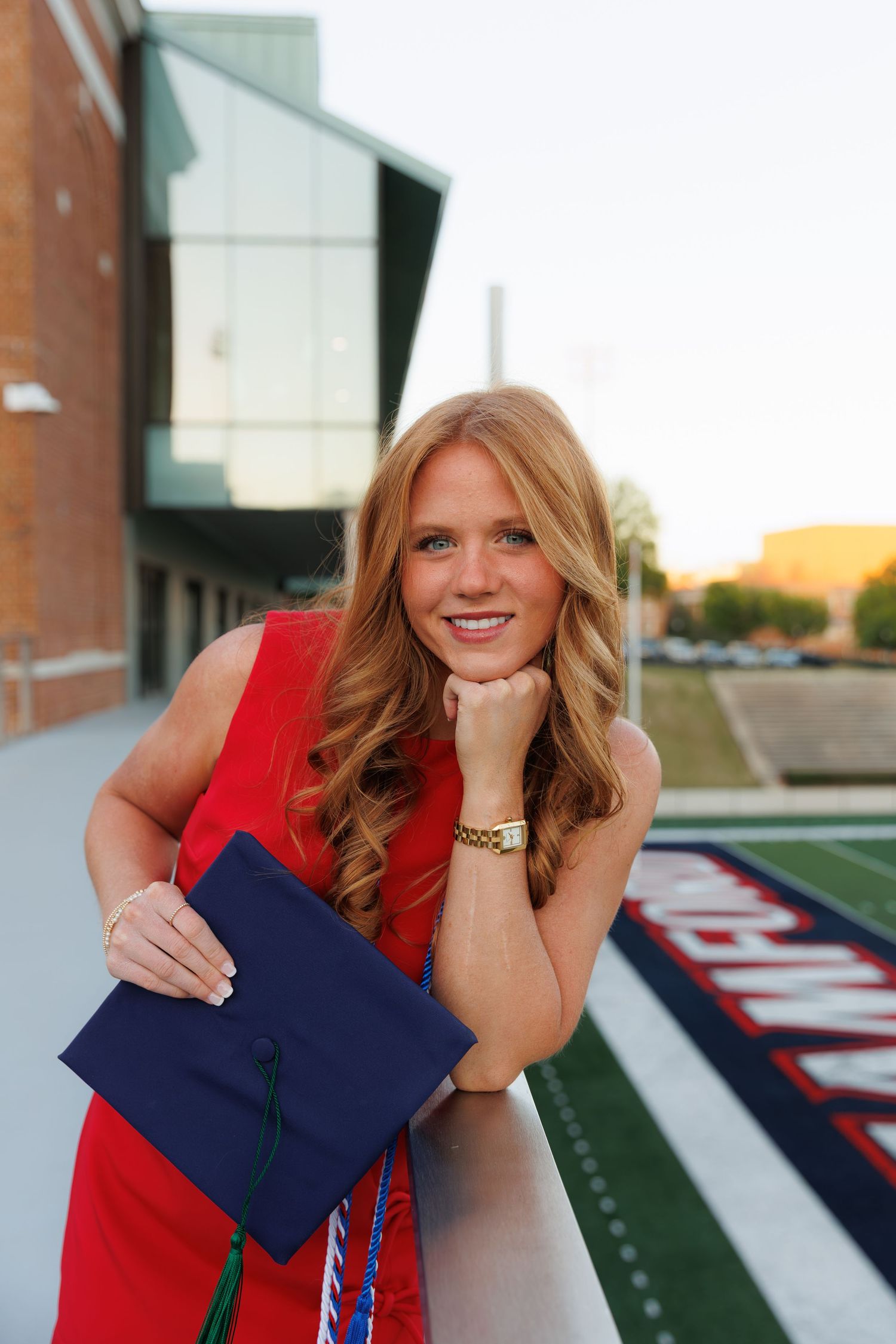 Smiling young woman in red dress holding navy graduation cap, leaning on railing at college stadium during golden hour.