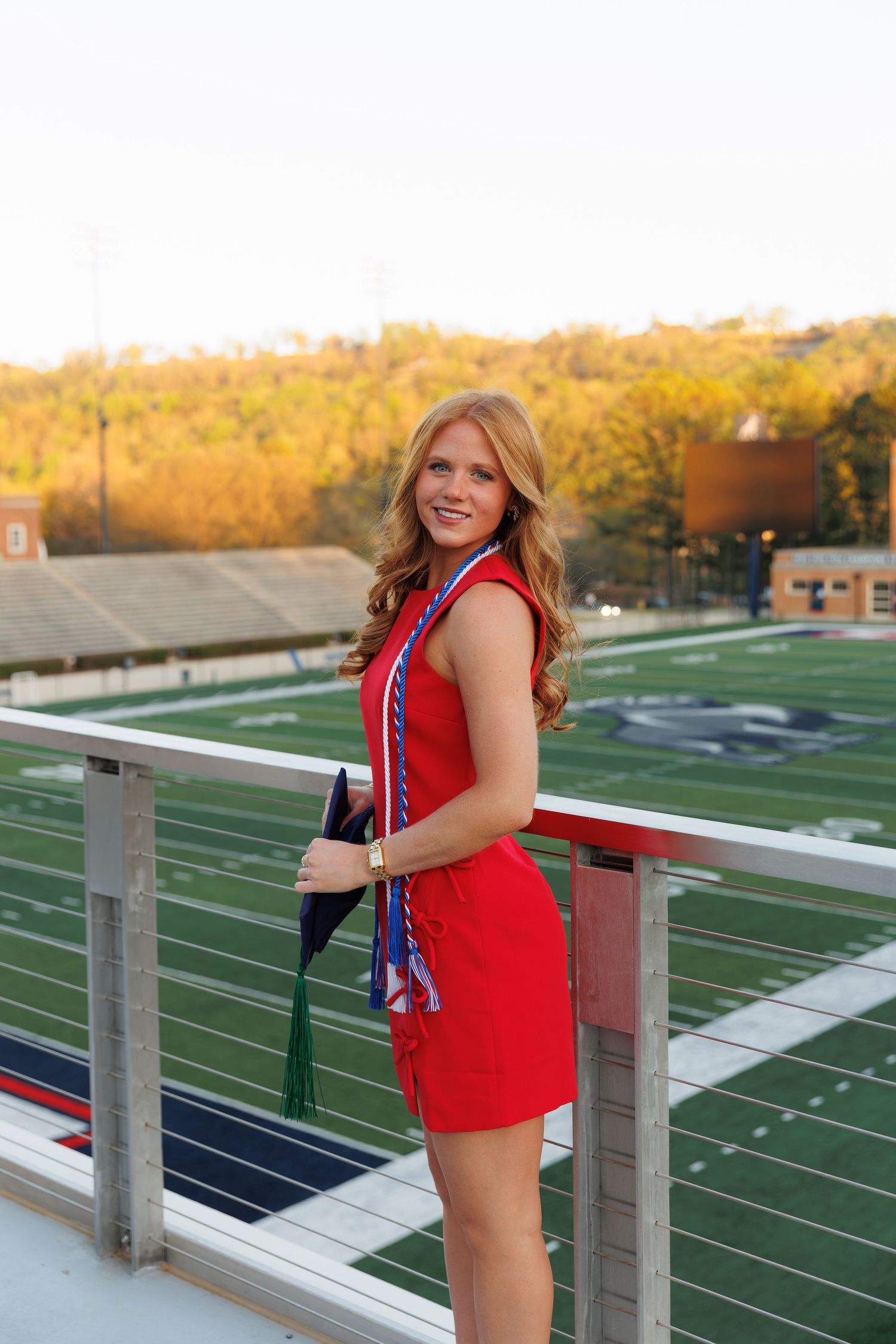 Smiling graduate in red dress with honor cords poses on stadium overlook at sunset.