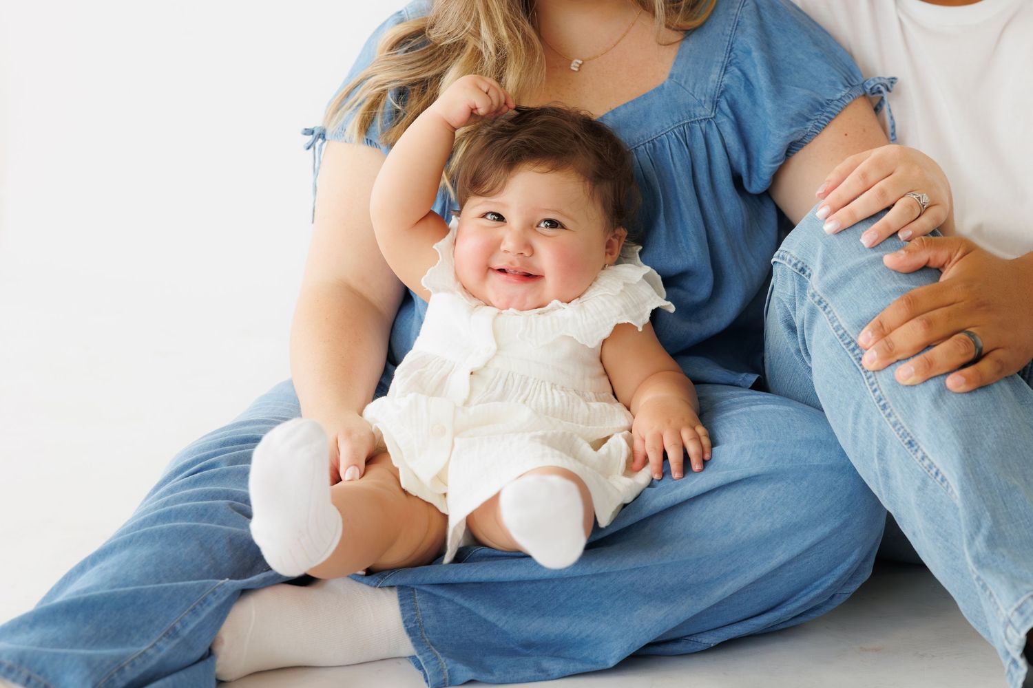 Smiling baby in white dress sitting between two adults in blue jeans and denim tops on white background.