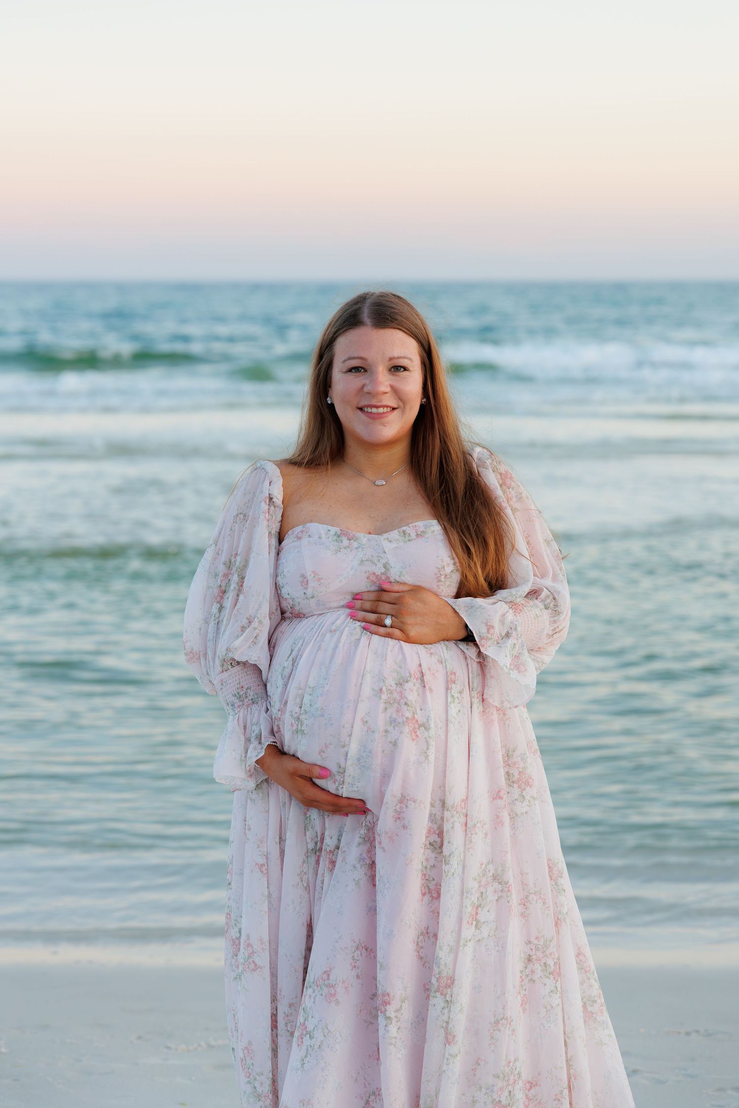 Pregnant woman in floral pink maternity gown smiling on beach at sunset, cradling baby bump near ocean waves.