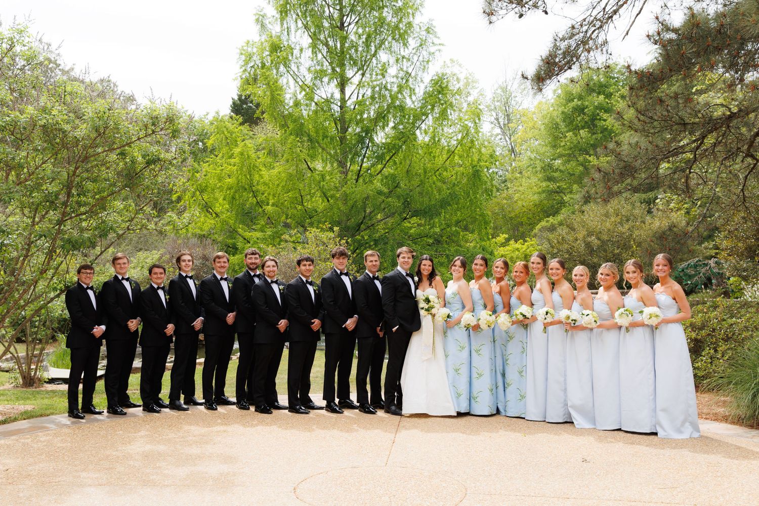 Wedding party posing outdoors, groomsmen in black tuxedos and bridesmaids in light blue gowns, with lush green trees behind.