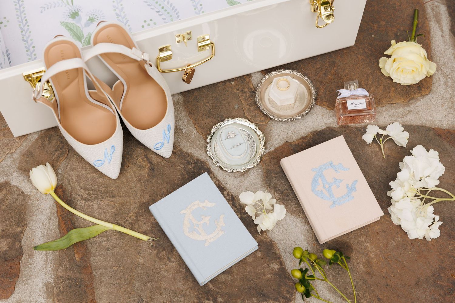 Elegant wedding flat lay featuring white heels, blue stationery, perfume, candles, and white flowers on a stone surface.