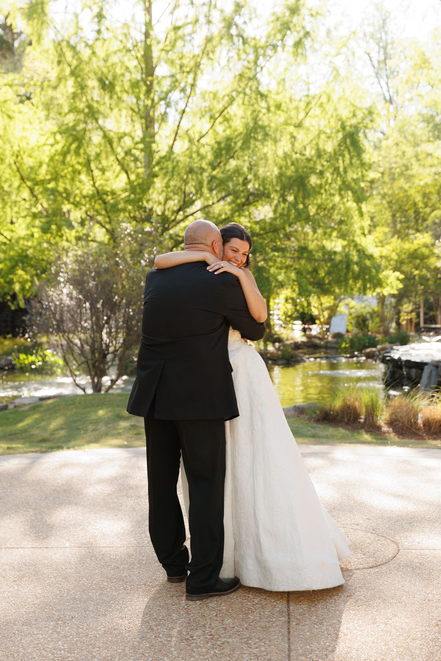 Newlyweds sharing a tender embrace in a sunlit garden, bride in white gown hugging groom in black suit.