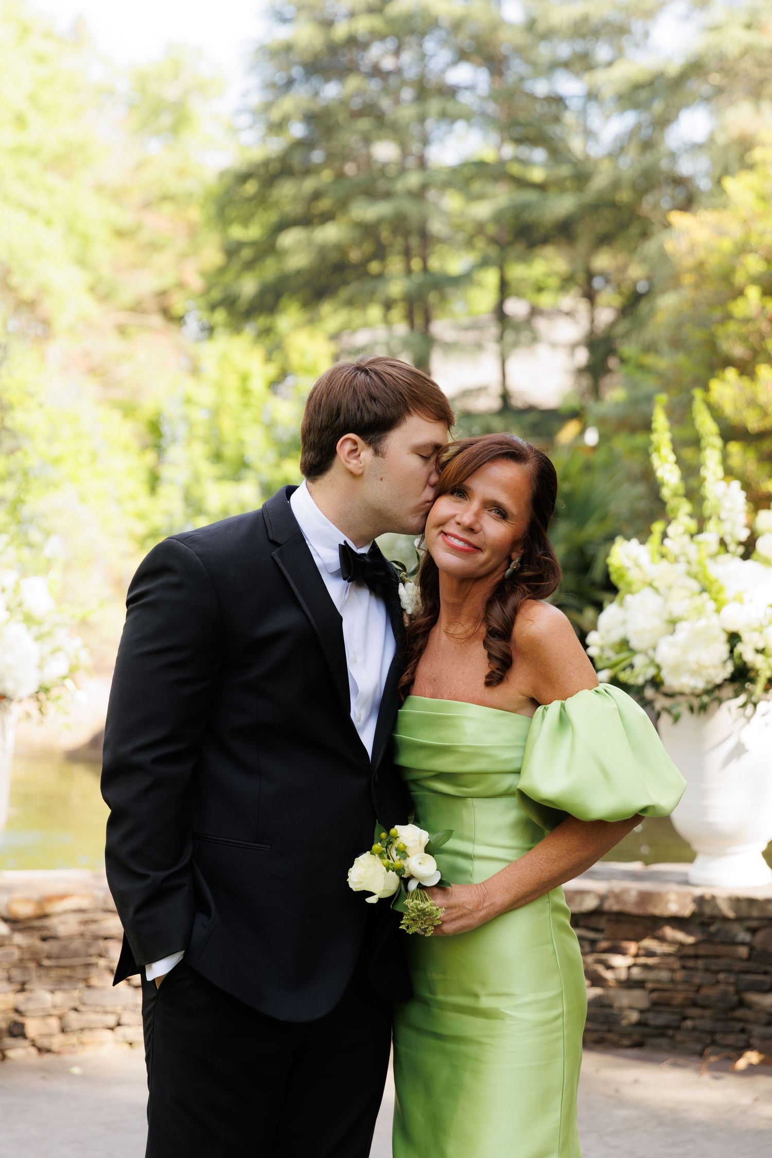 Groom in black tuxedo kisses woman in green off-shoulder gown at outdoor wedding ceremony.