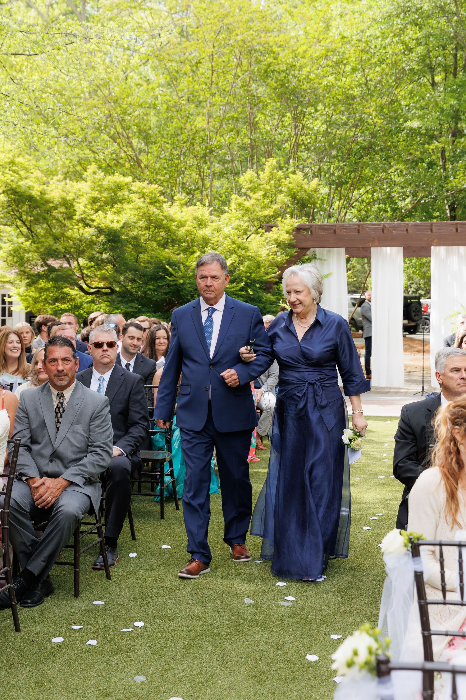 A man in a navy suit escorts an elderly woman in a navy dress down the aisle at an outdoor wedding ceremony.