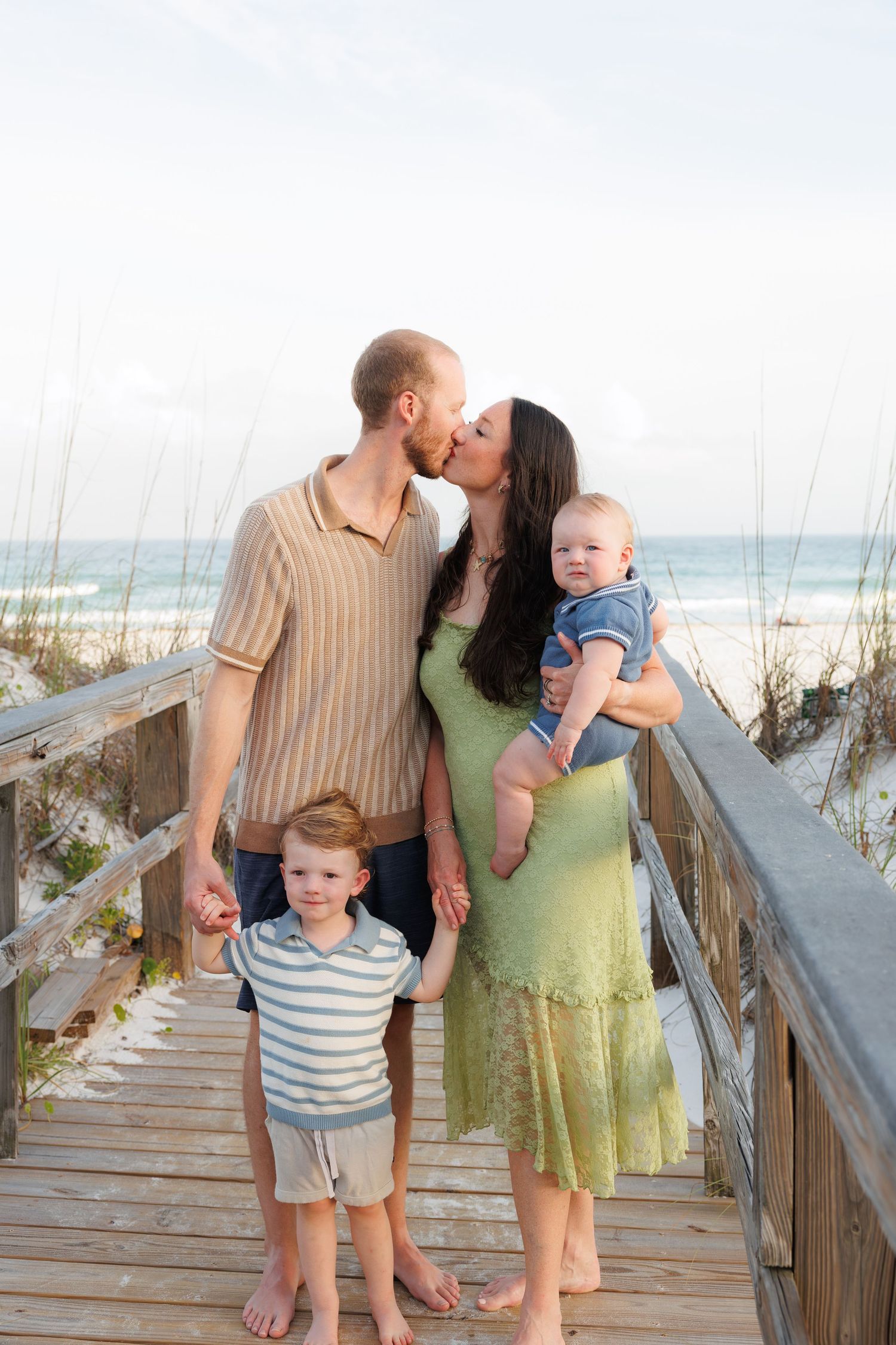 Family of four on beach boardwalk, parents kissing while mom holds baby and dad holds toddler's hand.
