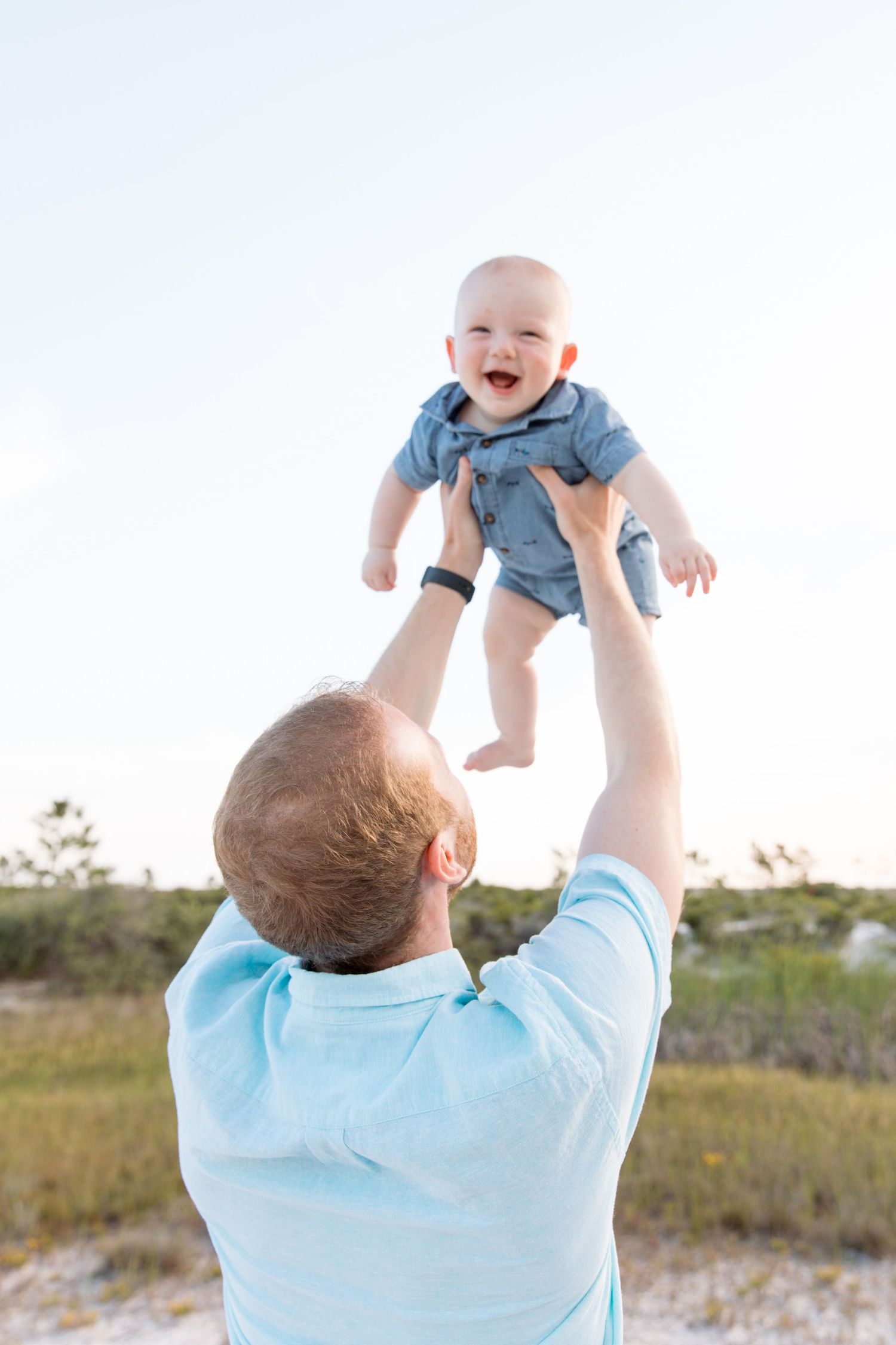 Father joyfully lifting laughing baby in blue onesie outdoors against bright sky in grassy field.