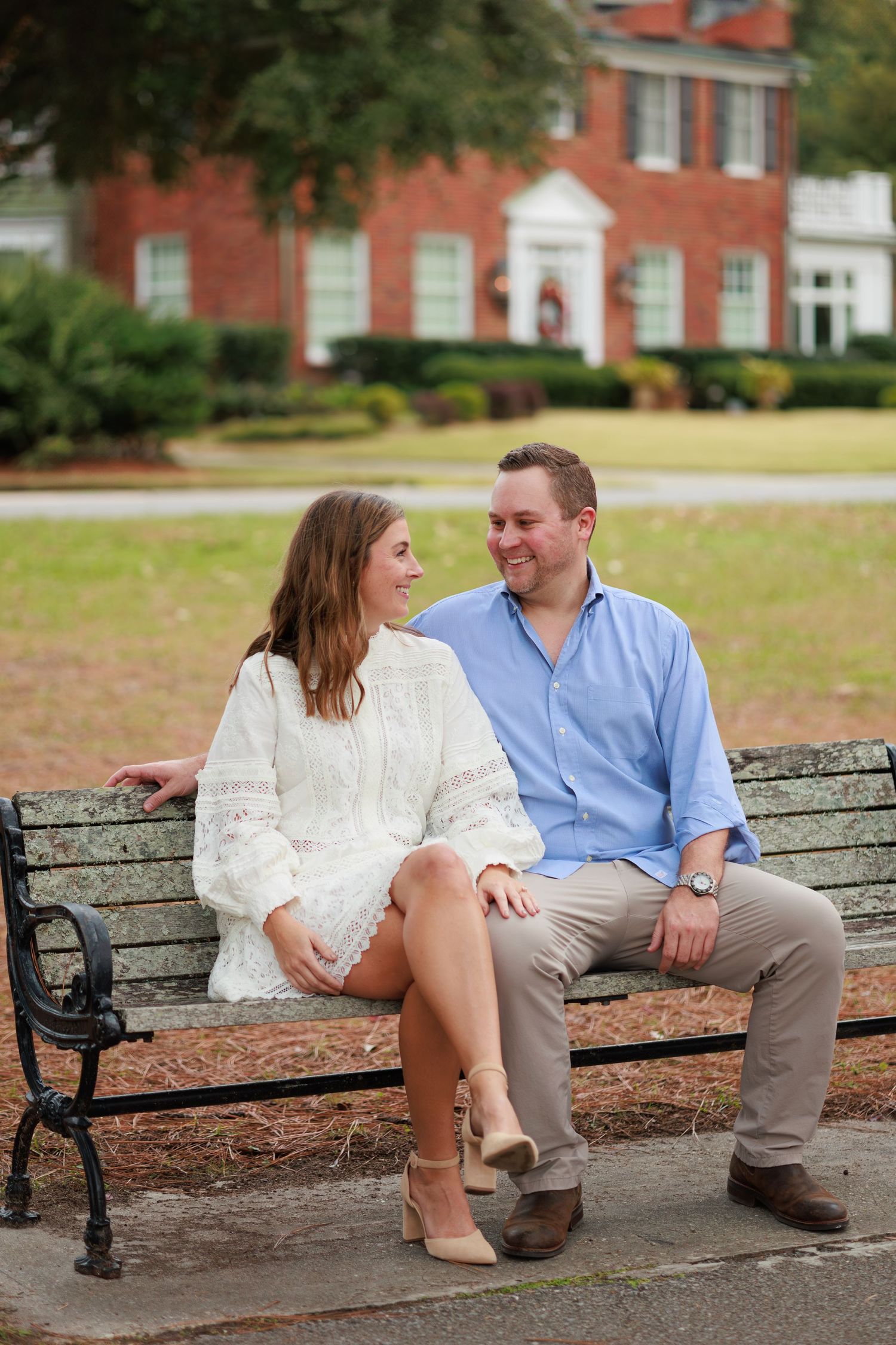A couple sits together on an ornate park bench in front of a red brick building on a fall day.
