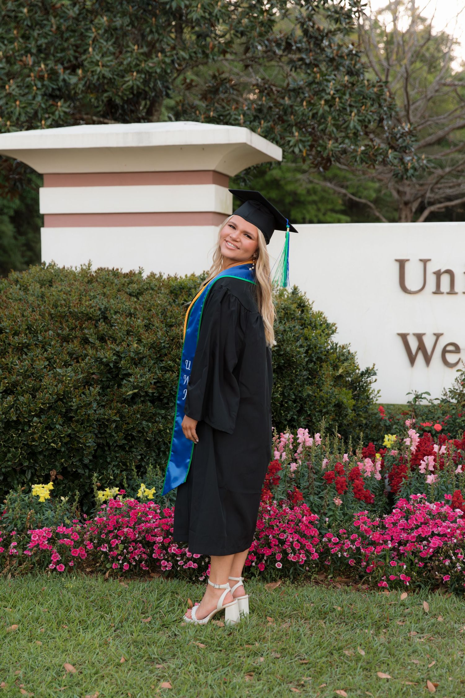 Graduate in cap and gown poses near pink azalea bushes and white stone wall during outdoor graduation photoshoot.