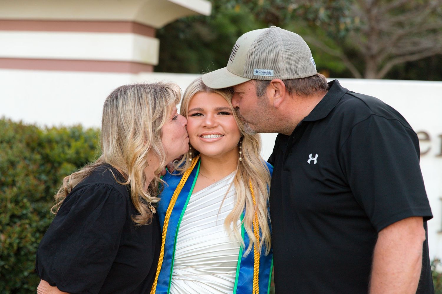 Parents kiss their graduate wearing blue sash and white graduation gown during outdoor celebration.