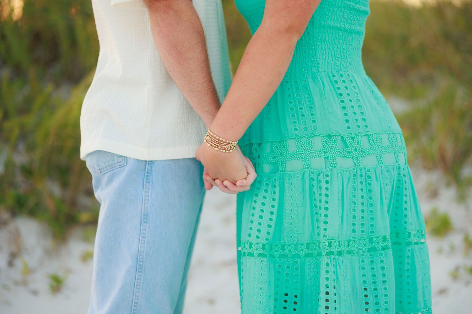 A couple in coordinated white and turquoise outfits holds hands during a romantic outdoor photoshoot at sunset.