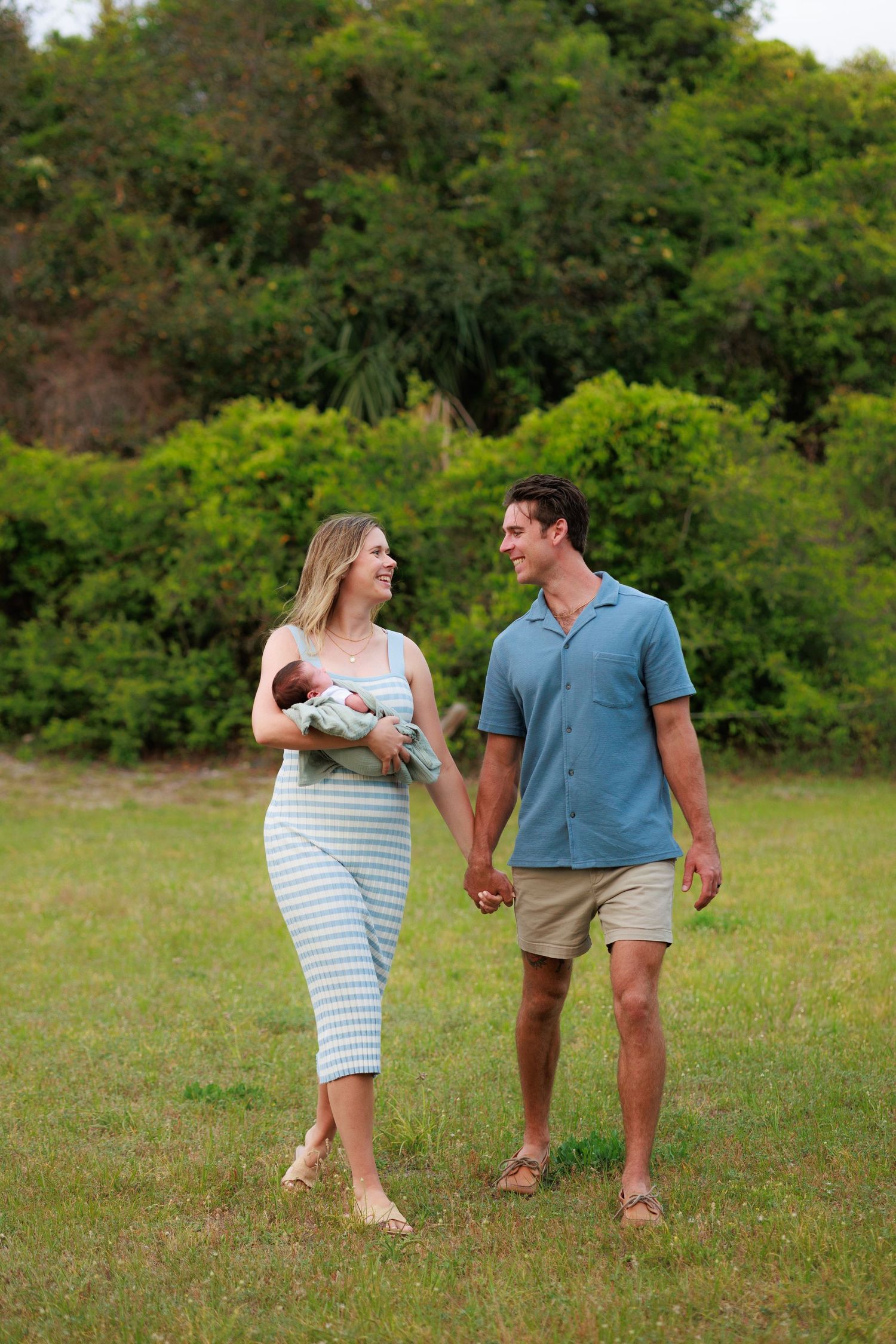 A couple walks hand in hand across a grassy field with lush green trees in the background.