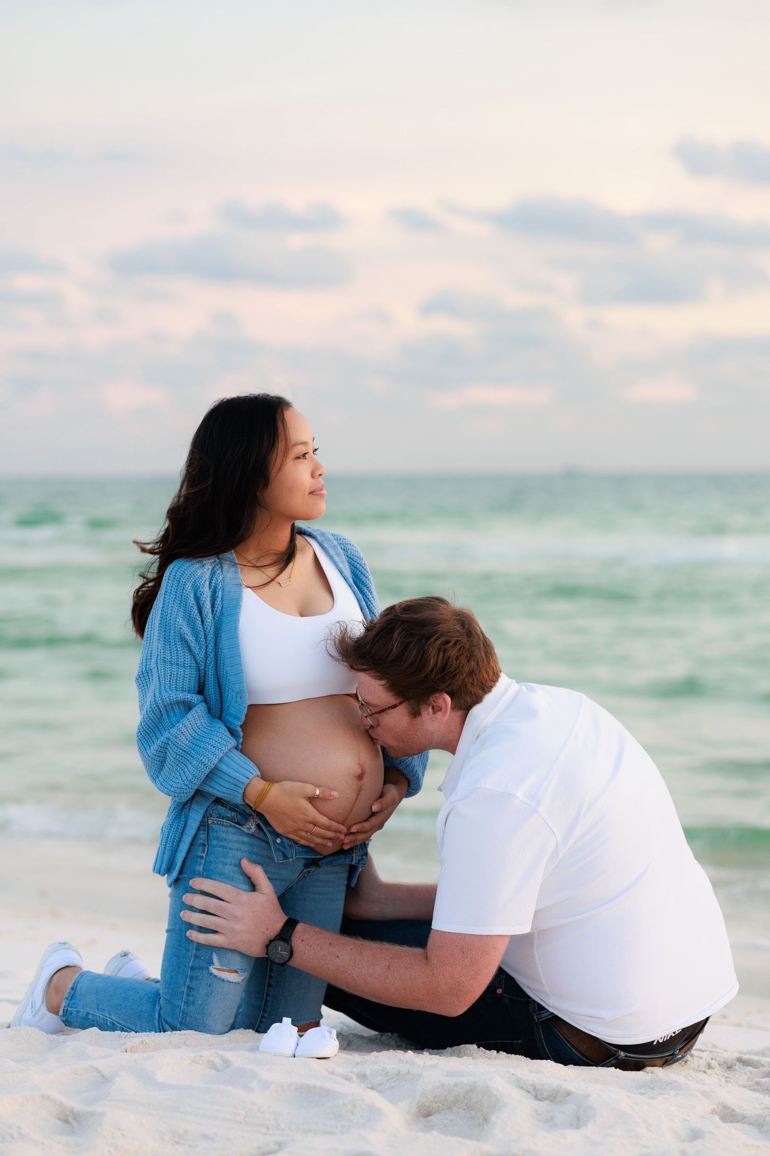 Couple taking romantic maternity photos on a sandy beach at sunset with ocean waves in the background.