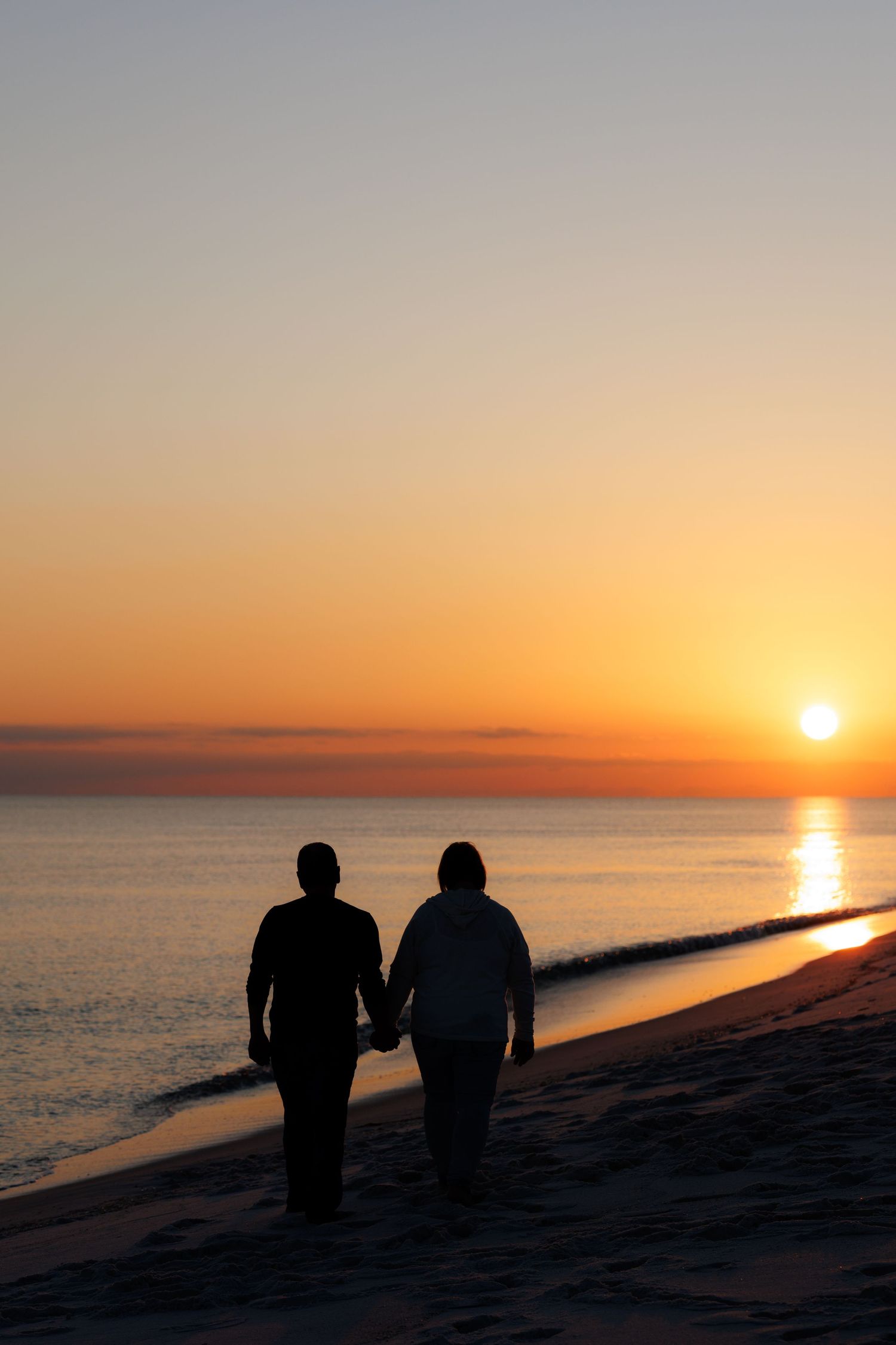 Silhouettes of two people walking along a beach at sunset with the orange sun reflecting on the ocean.