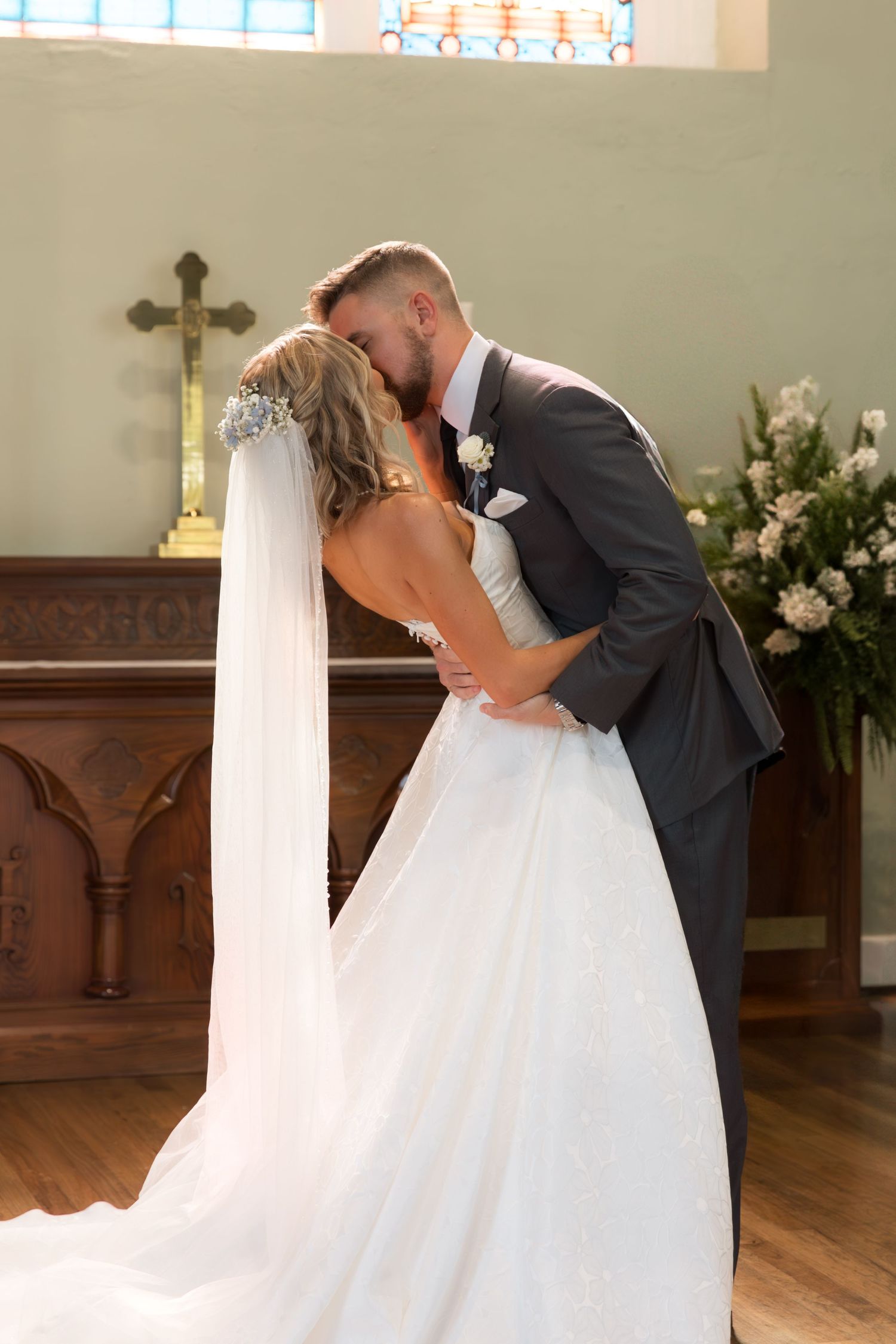 A romantic first kiss between newlyweds in a church with a cross and floral decorations in the background.
