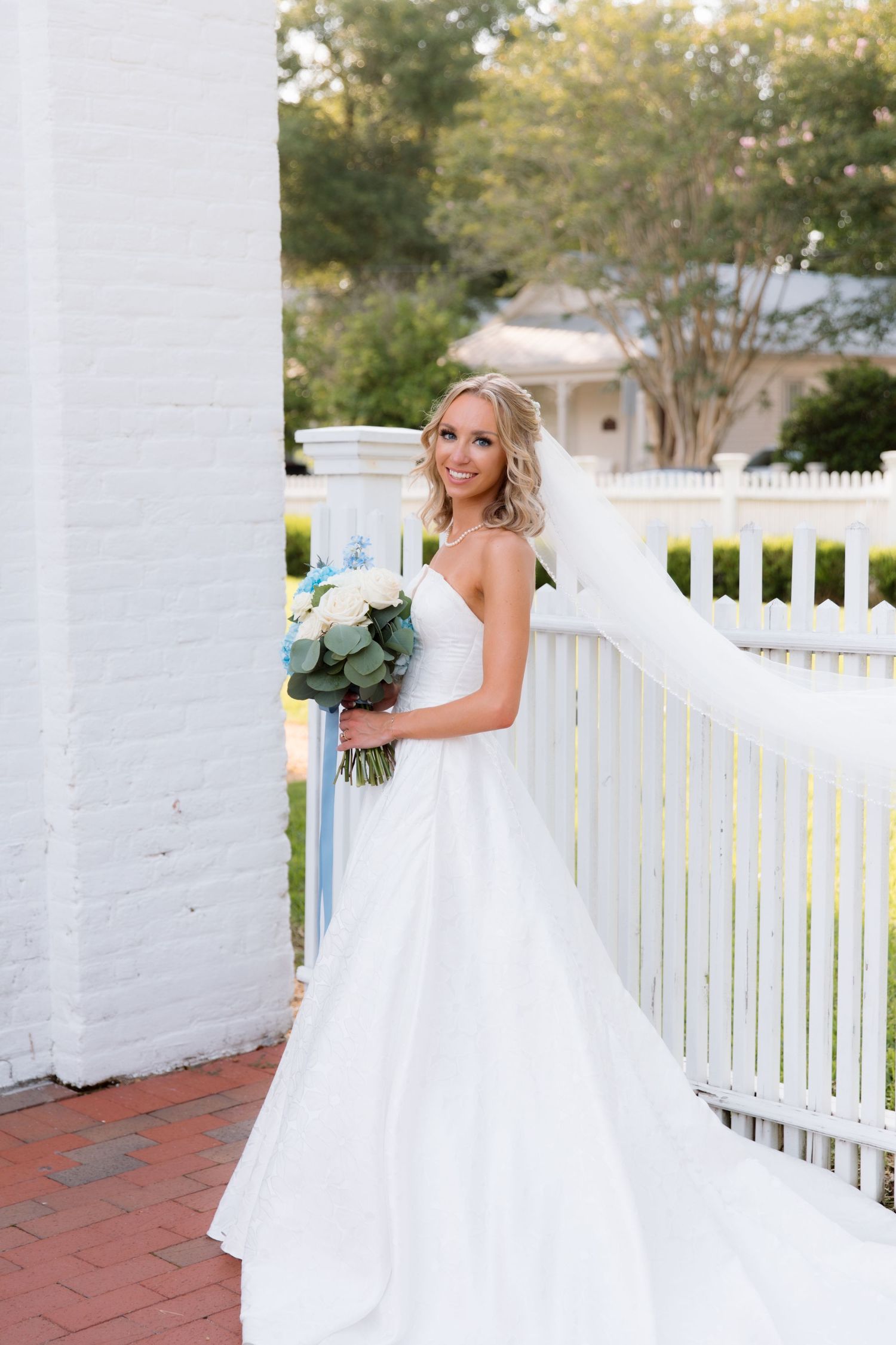 A bride poses in a classic white wedding gown against a white picket fence.