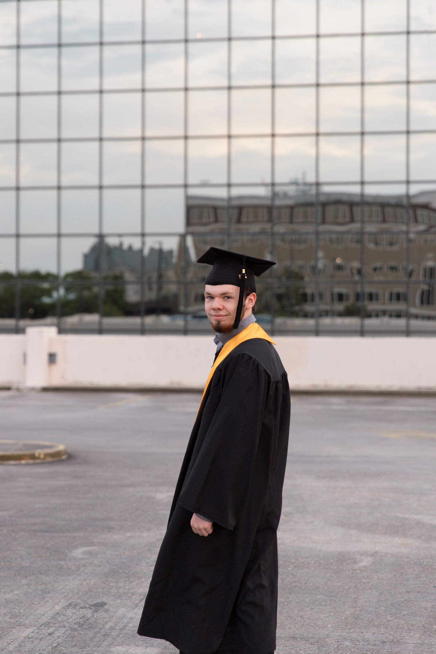 Graduate in black graduation robe and cap with gold sash standing on urban rooftop.
