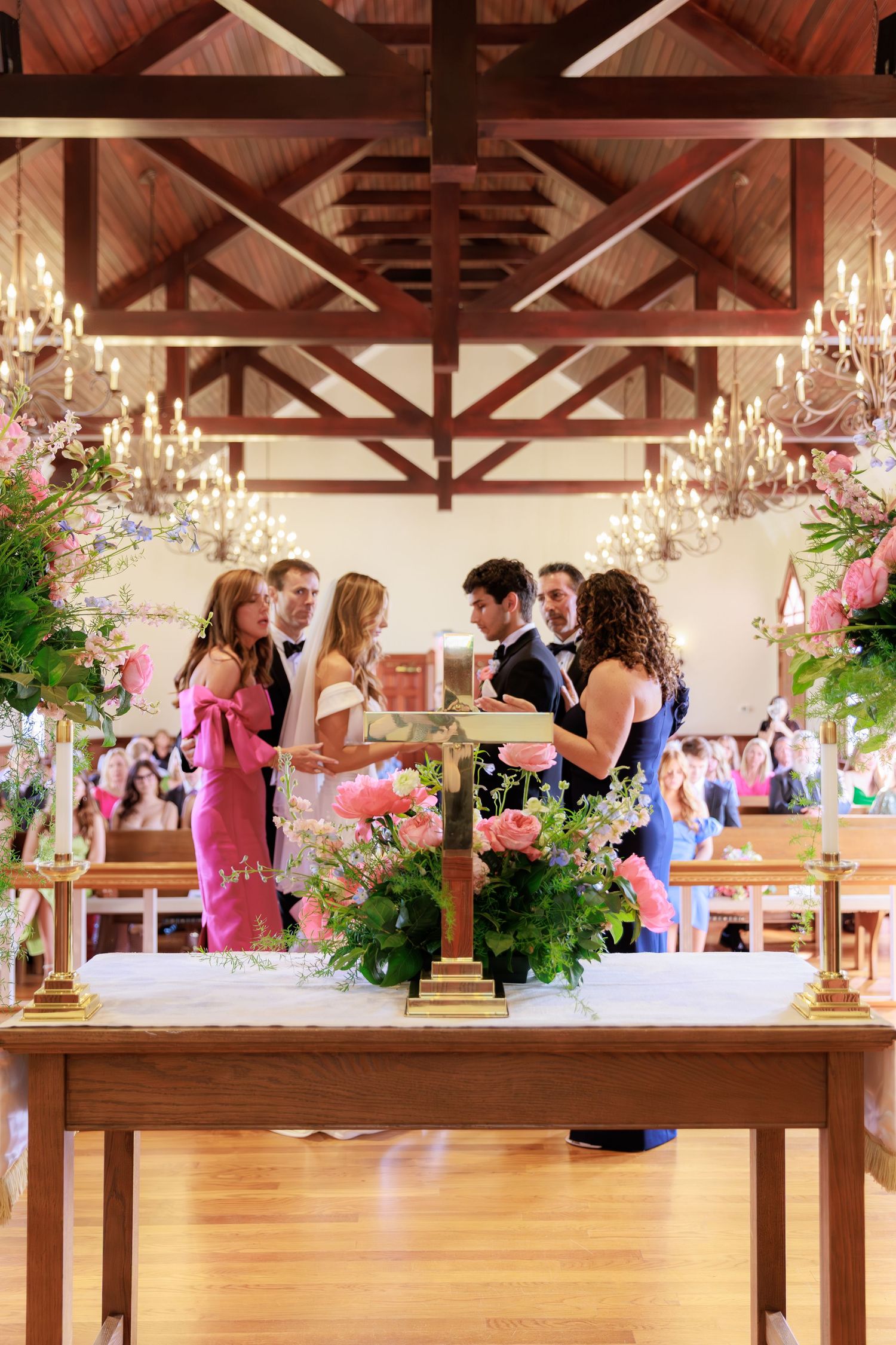 A group gathers around a decorated table with floral arrangements under wooden beams and crystal chandeliers.