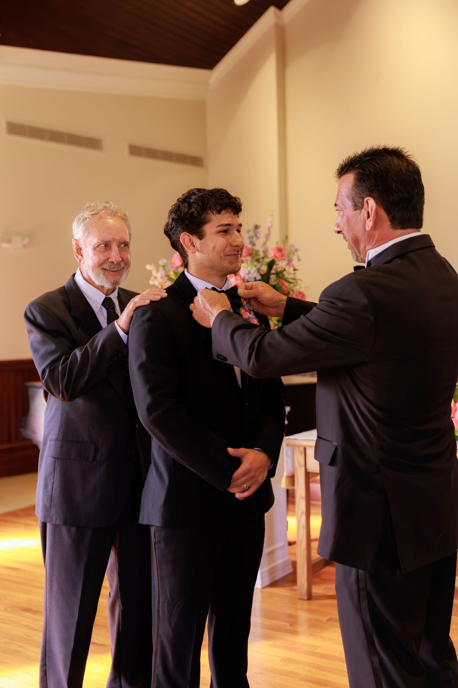 Two men in suits help adjust another man's bowtie during a formal event in a room with hardwood floors.