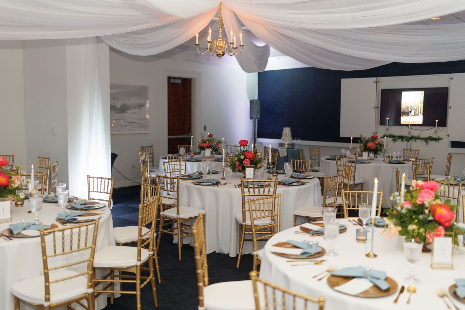 An elegant wedding reception hall with white draped ceiling, gold chiavari chairs and coral floral centerpieces on round tables.