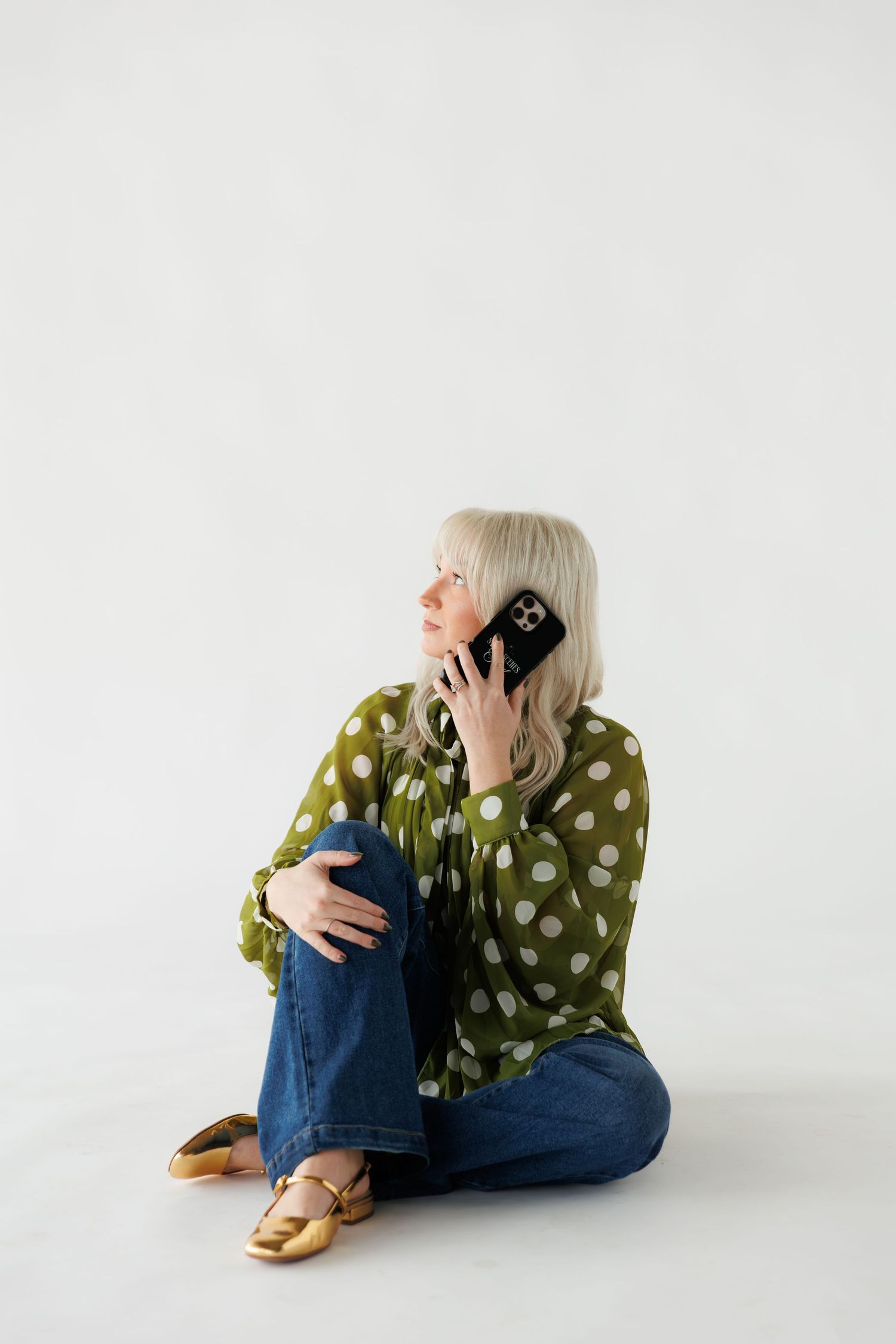 Older woman with silver hair sitting on floor in green polka dot blouse and jeans, talking on smartphone.