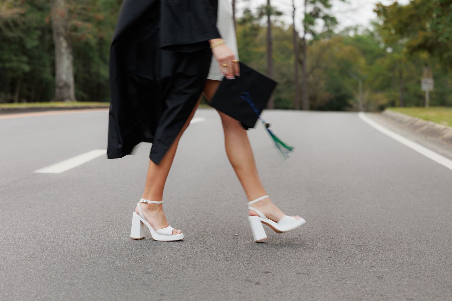 Graduate in black gown and white block heels holding cap walks on road during graduation photo shoot.