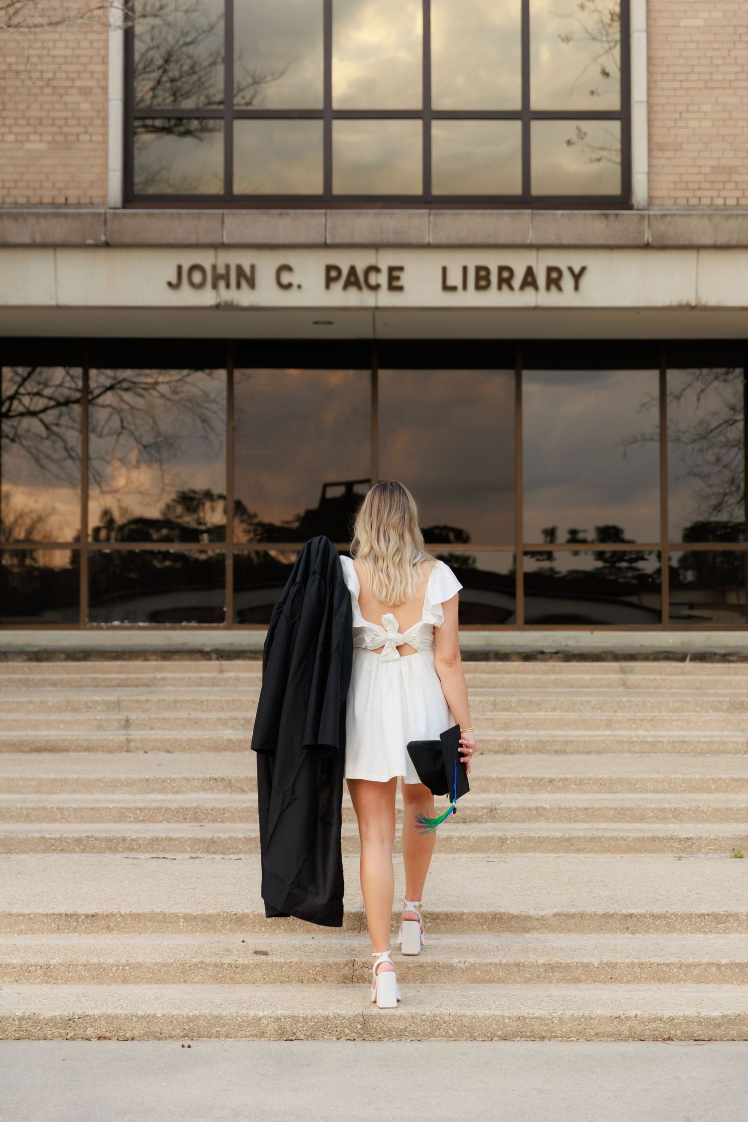Blonde woman in white dress holding graduation gown walks up steps to John C. Pace Library.