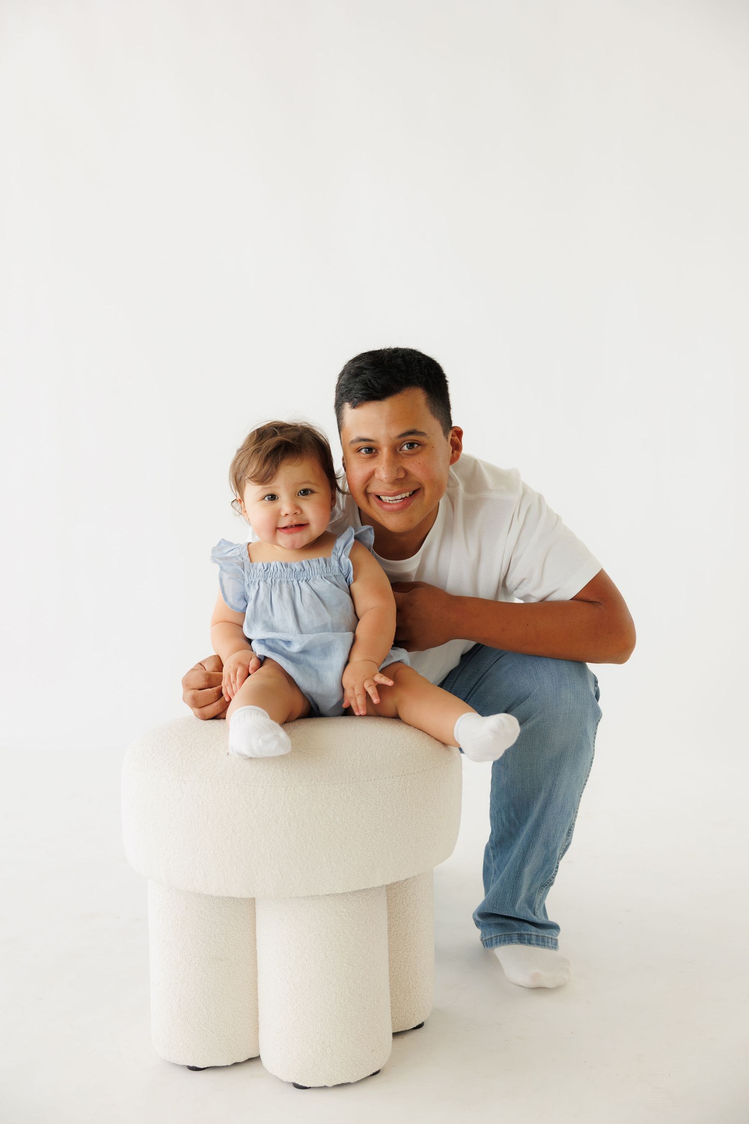 A smiling father crouches beside his baby girl in a blue dress, seated on a white stool, in a bright studio.