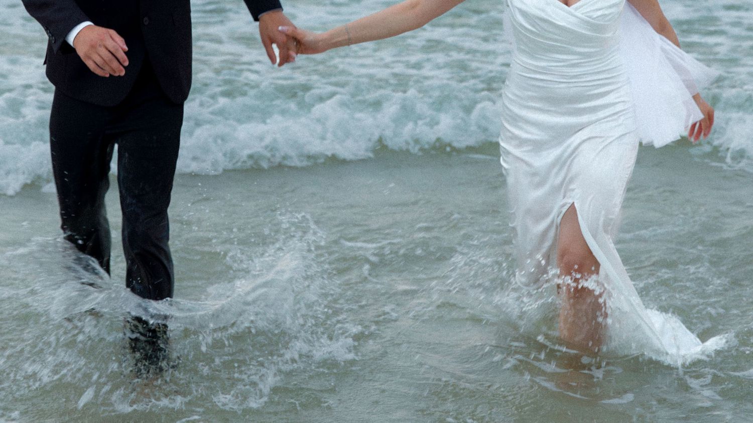 Newlyweds splash in ocean waves while holding hands.