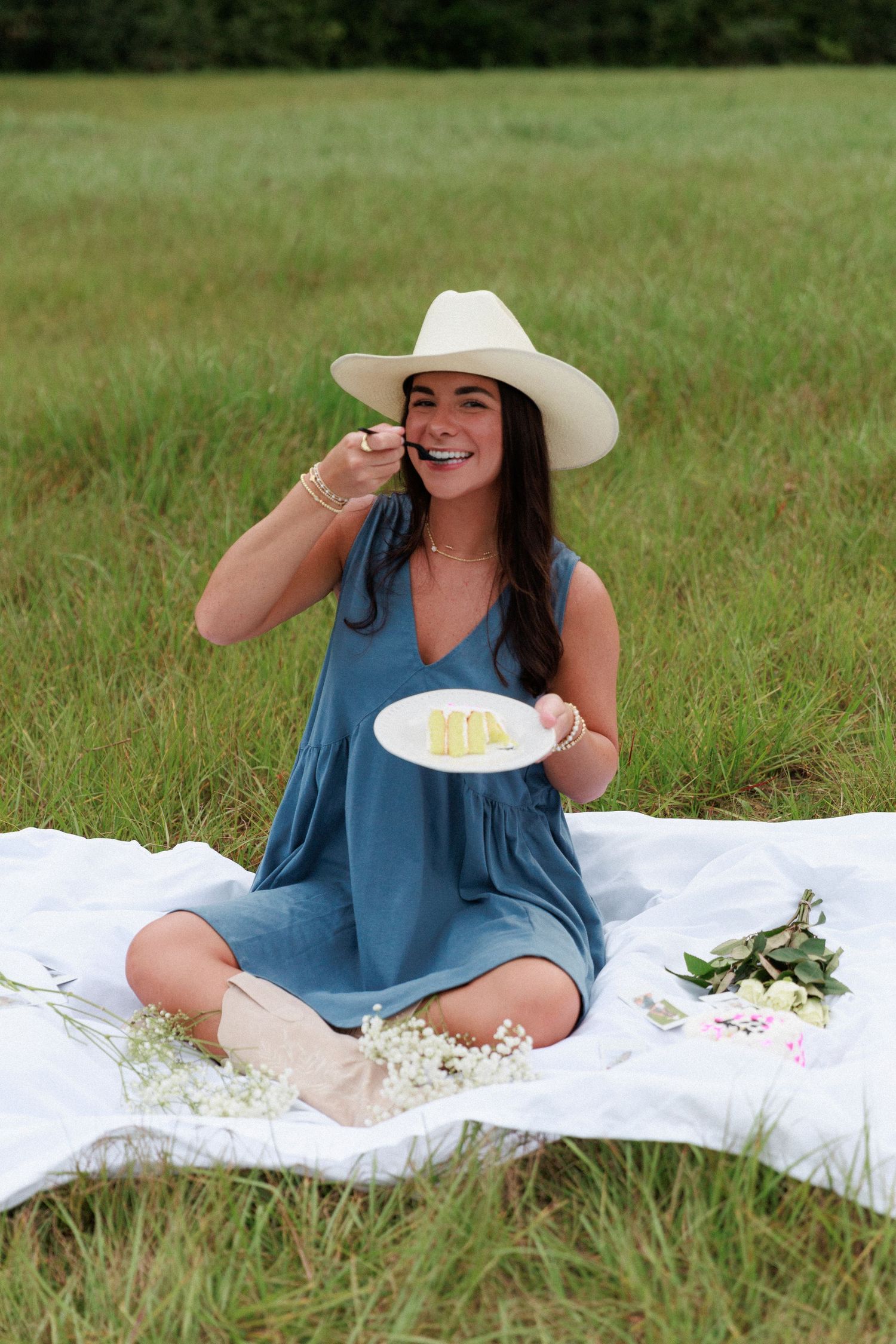 Person in blue dress and cowboy hat enjoying cake during outdoor picnic.