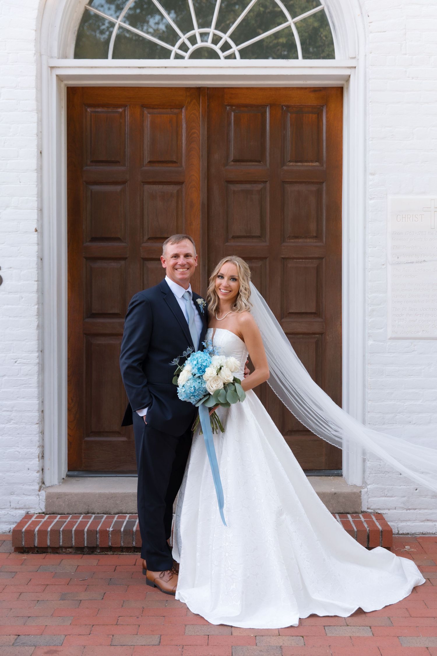Wedding couple poses elegantly in front of a classic wooden church door with a stunning white dress and blue bouquet.