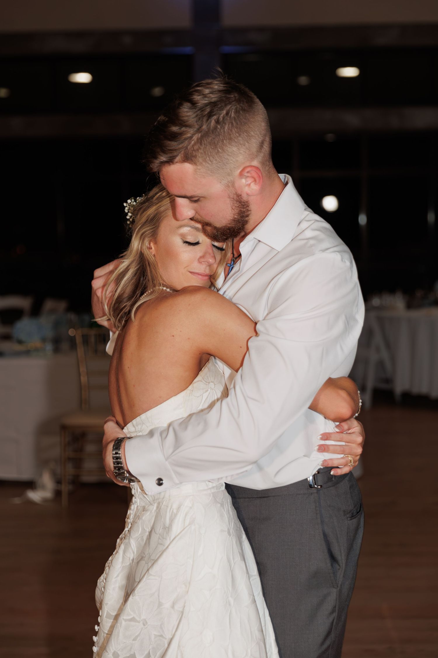 Couple embraces in tender moment during wedding dance, bride wearing white gown.
