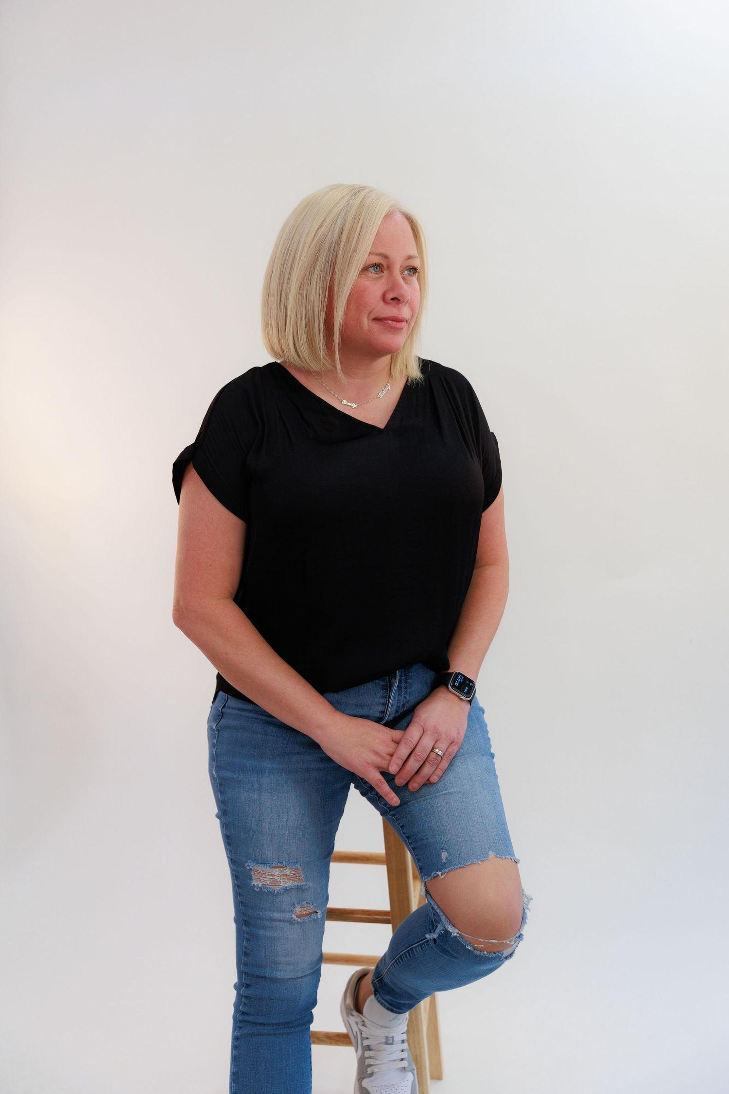 Casual studio portrait with black top and ripped jeans while resting foot on wooden stool.