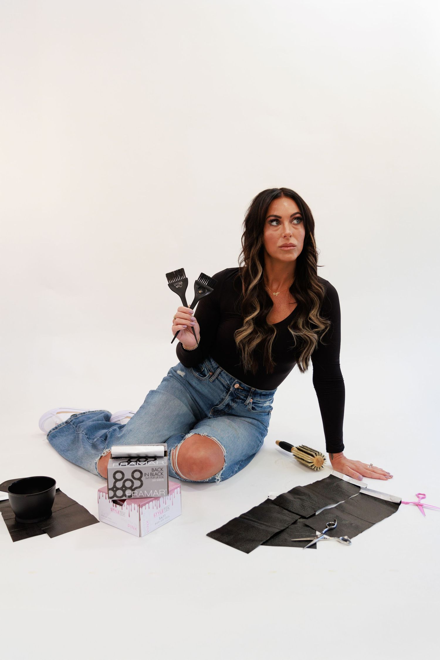 Beauty professional with styling tools sits on white floor surrounded by hair products.
