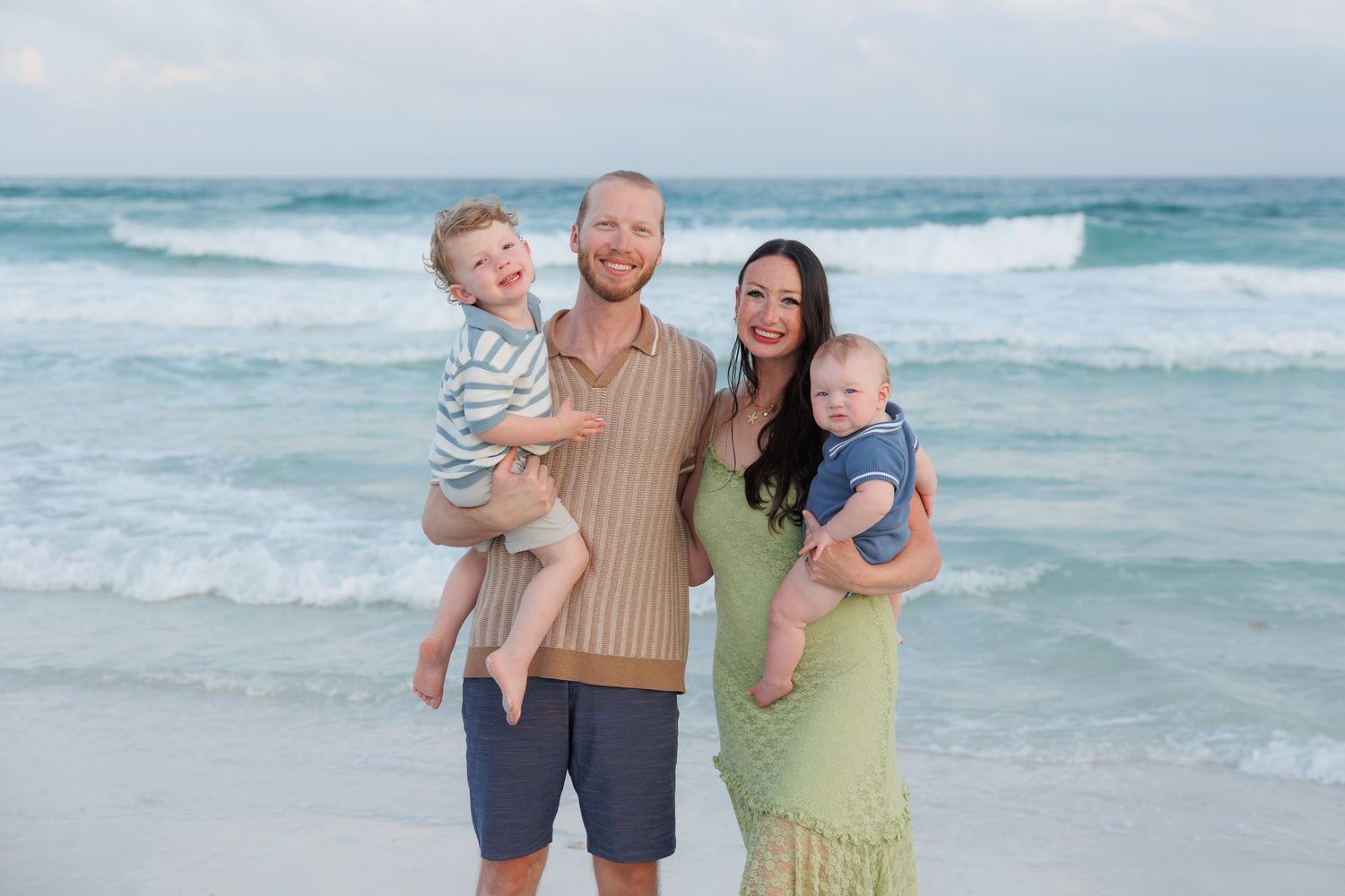 Smiling family of four with father holding two young boys standing on sunny white sand beach.