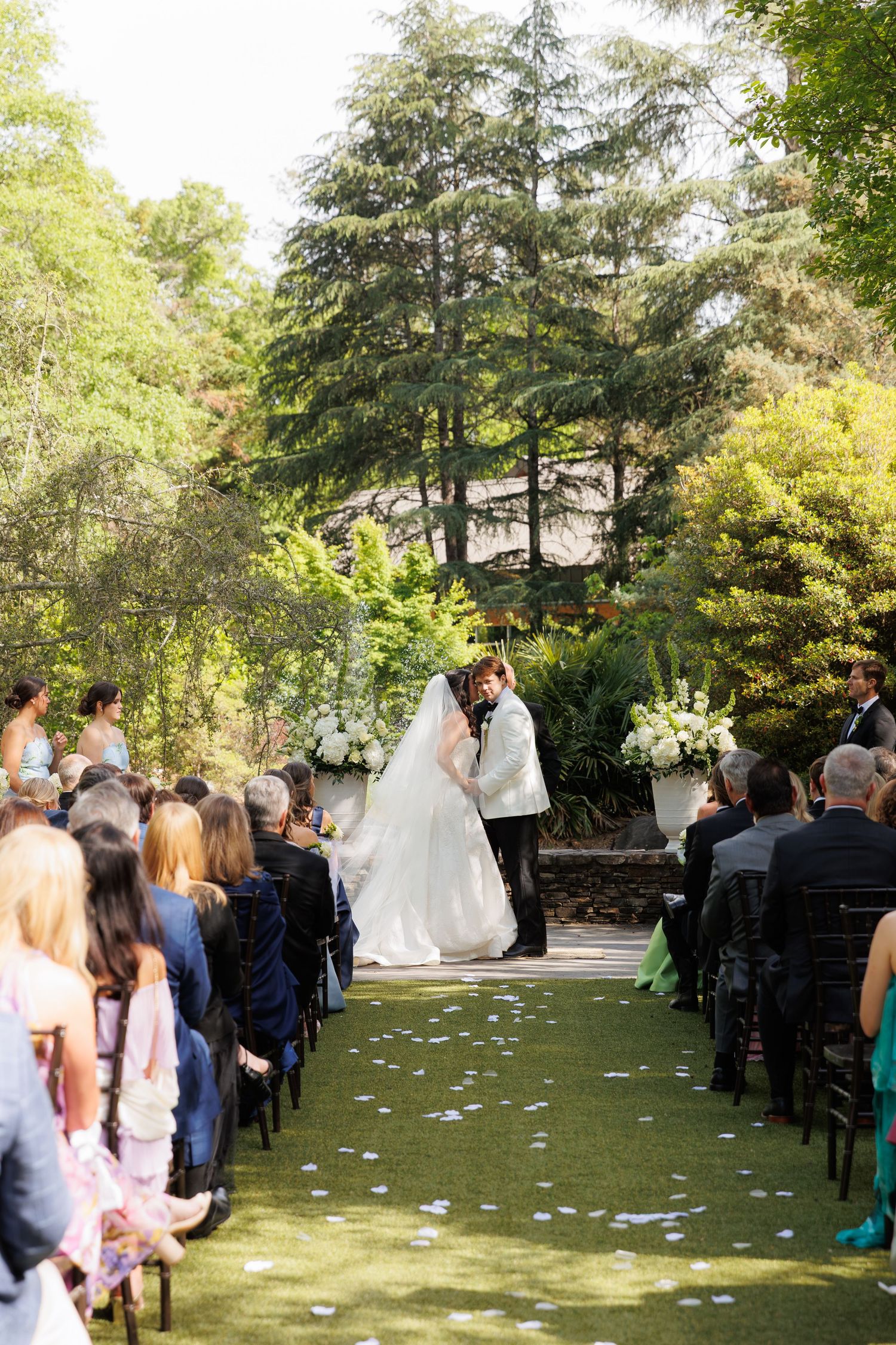 Bride and groom share first kiss at outdoor garden wedding ceremony with white floral arrangements and tree-lined aisle.