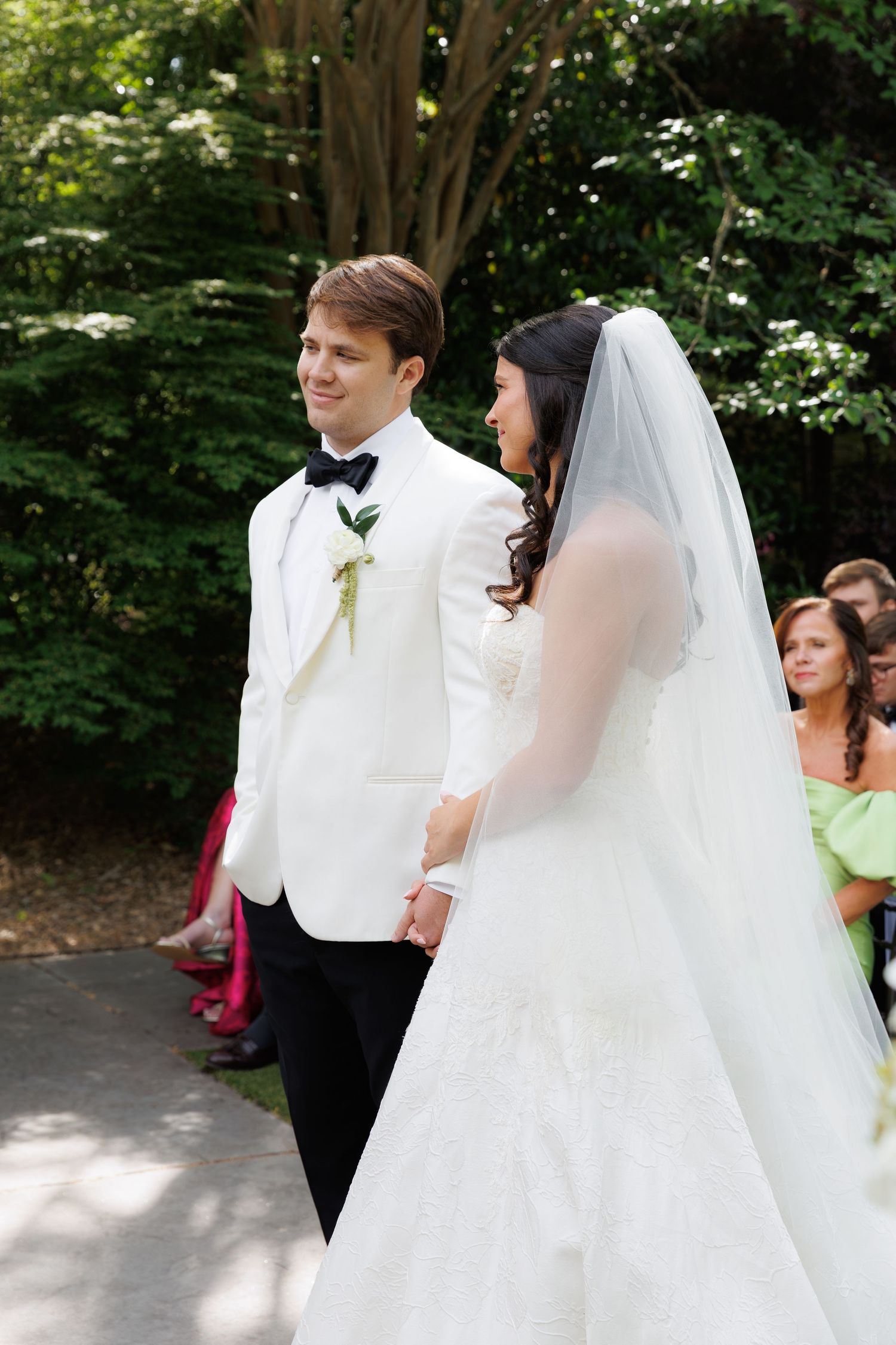 Bride and groom hold hands during outdoor wedding ceremony, groom wearing white tuxedo, bride in white gown with veil.