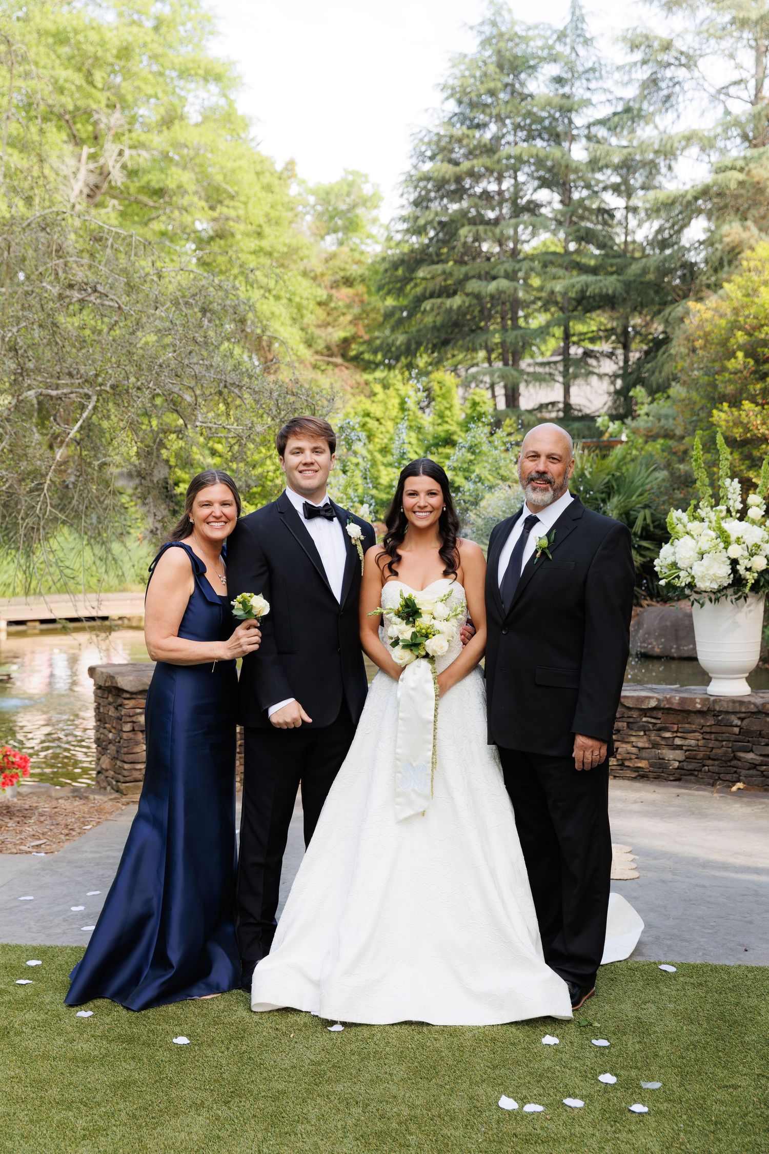 Wedding party of four posing outdoors, bride in white gown with bouquet, groom in tuxedo, guests in navy and black formal attire.